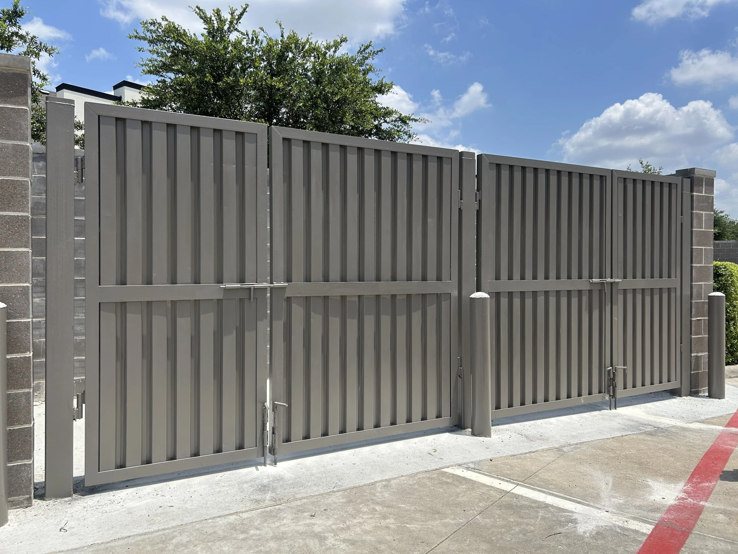 Gray metal double gate with vertical slats, set between brick posts, with a concrete driveway in front, under a partly cloudy blue sky.
