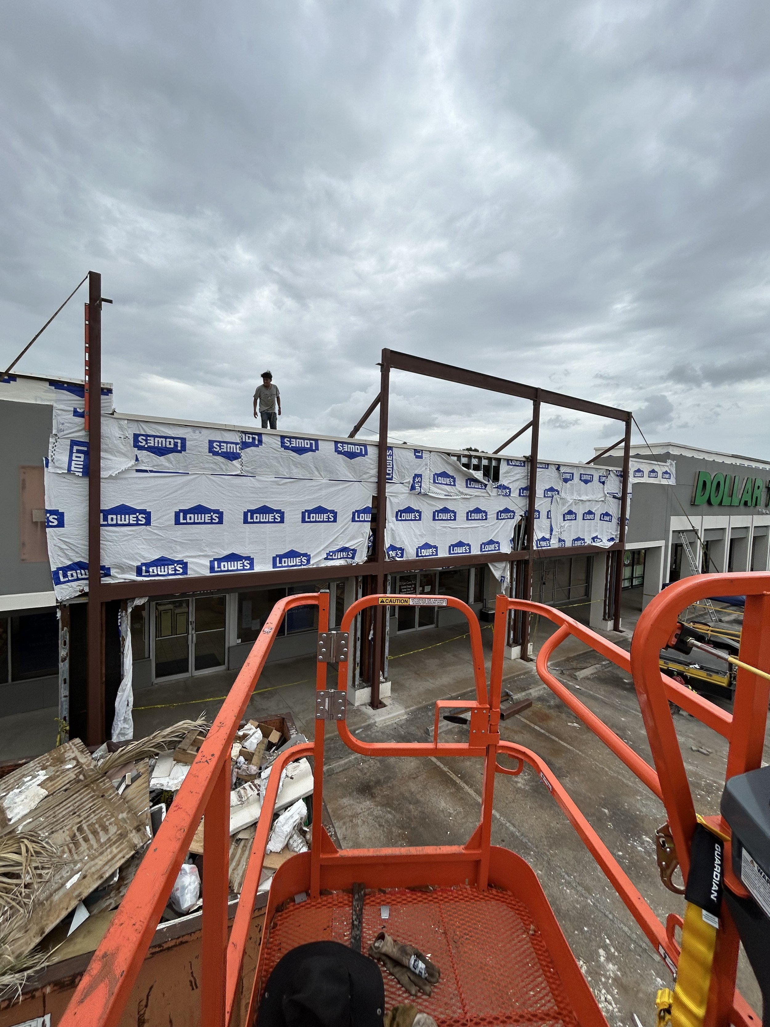 Construction worker on top of a building under renovation with scaffolding and Lowe's branded insulation, with a Dollar Tree store in the background, overcast sky.
