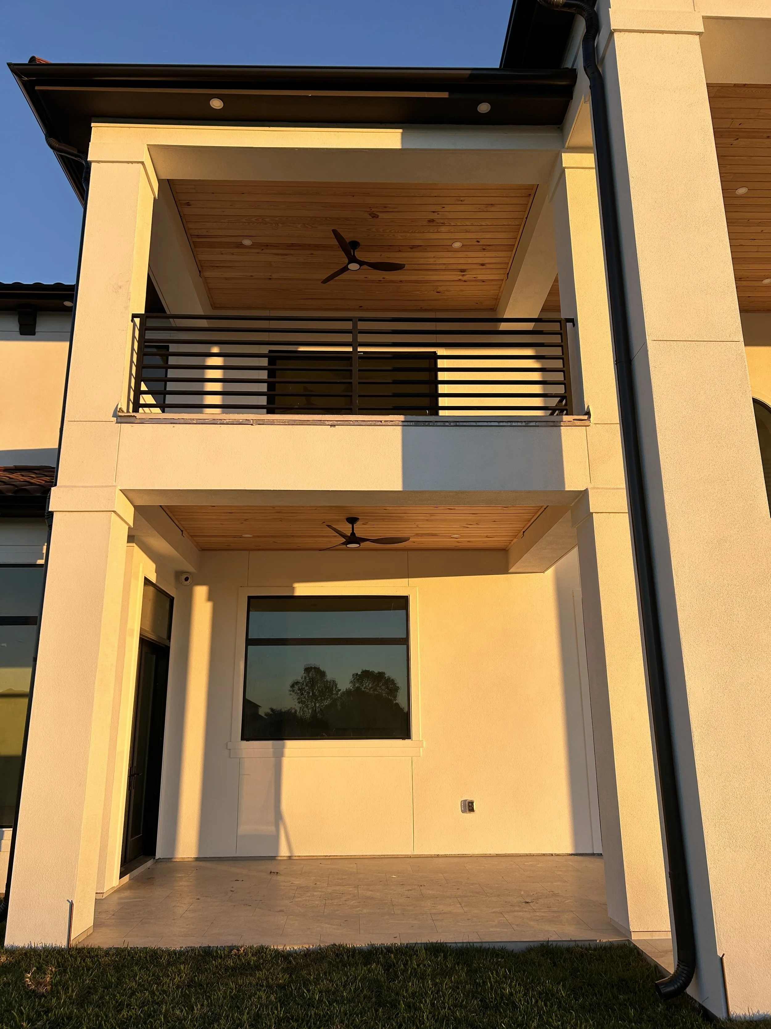 Front view of a modern two-story house with a balcony, ceiling fans, and large windows, during sunset.