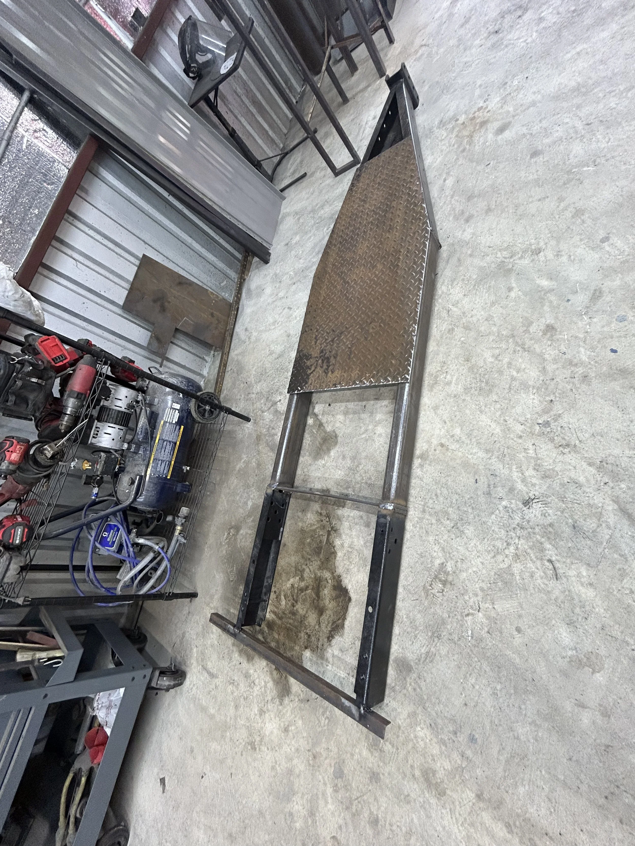 Metal frame with a slanted diamond plate section, lying on a concrete floor in a workshop.