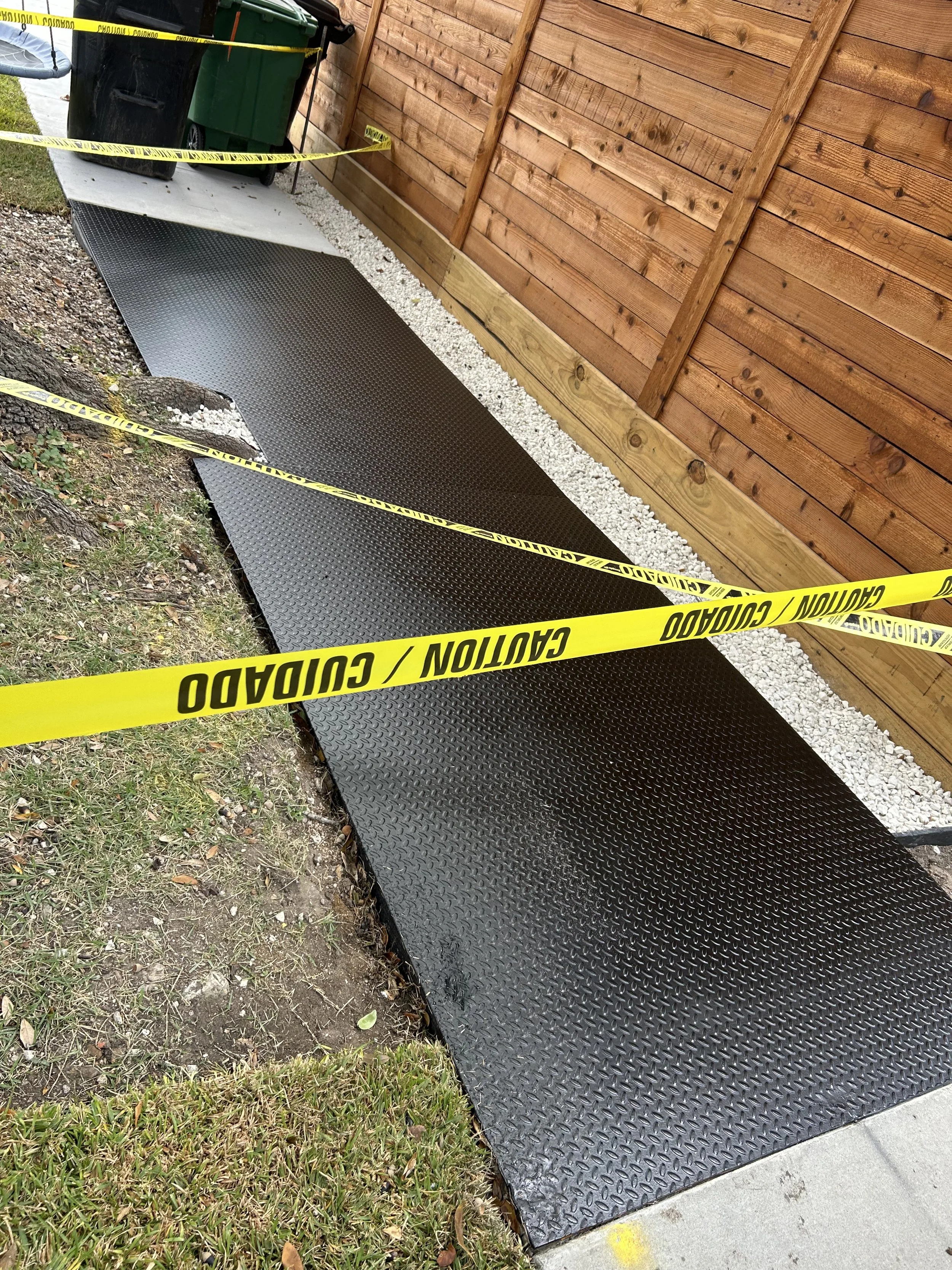Black metal ramp with yellow caution tape around it, situated outside next to a wooden fence and compost bins.