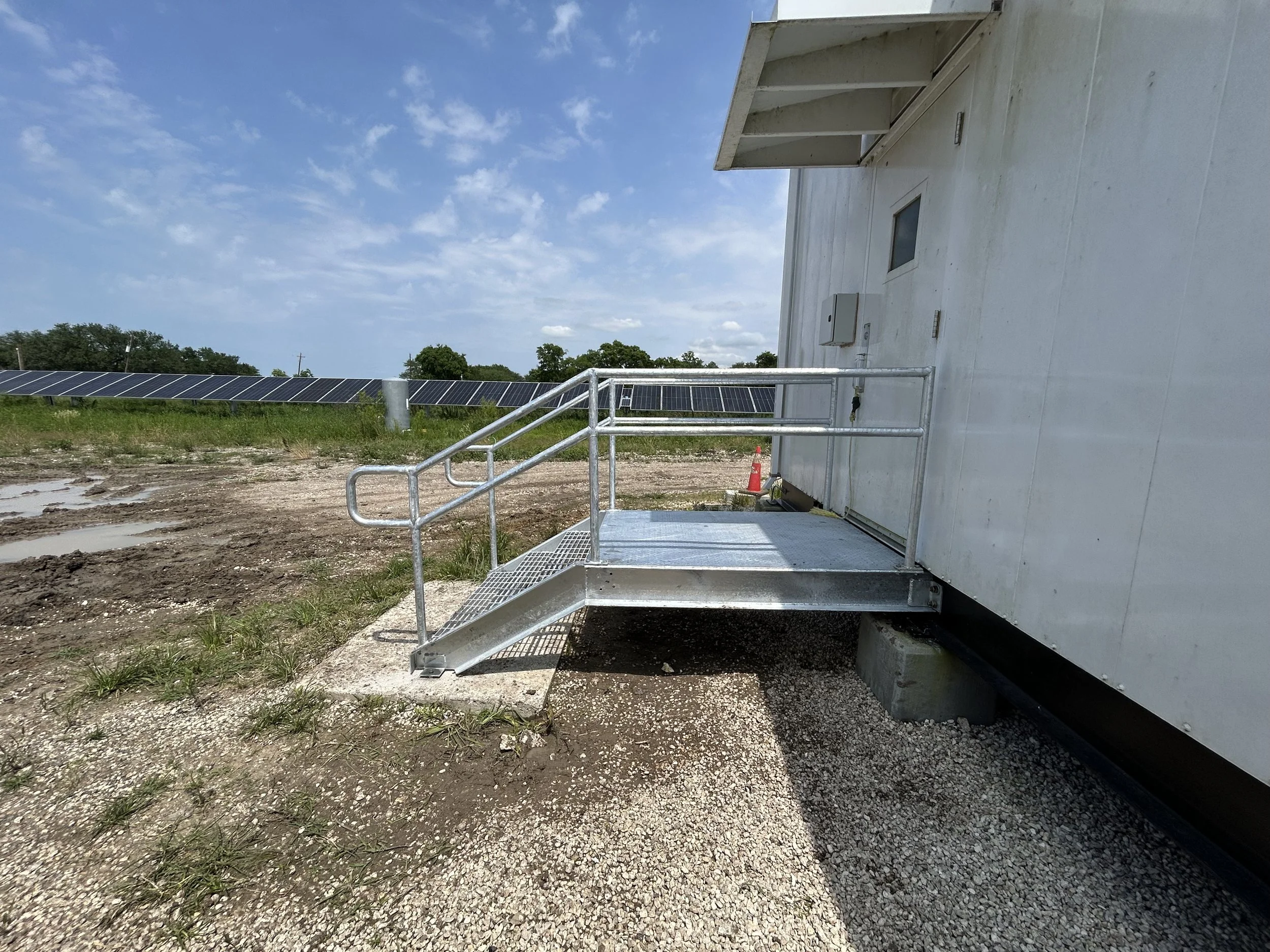 Metal wheelchair ramp outside a white building with solar panels in the background, grassy area, cloudy sky