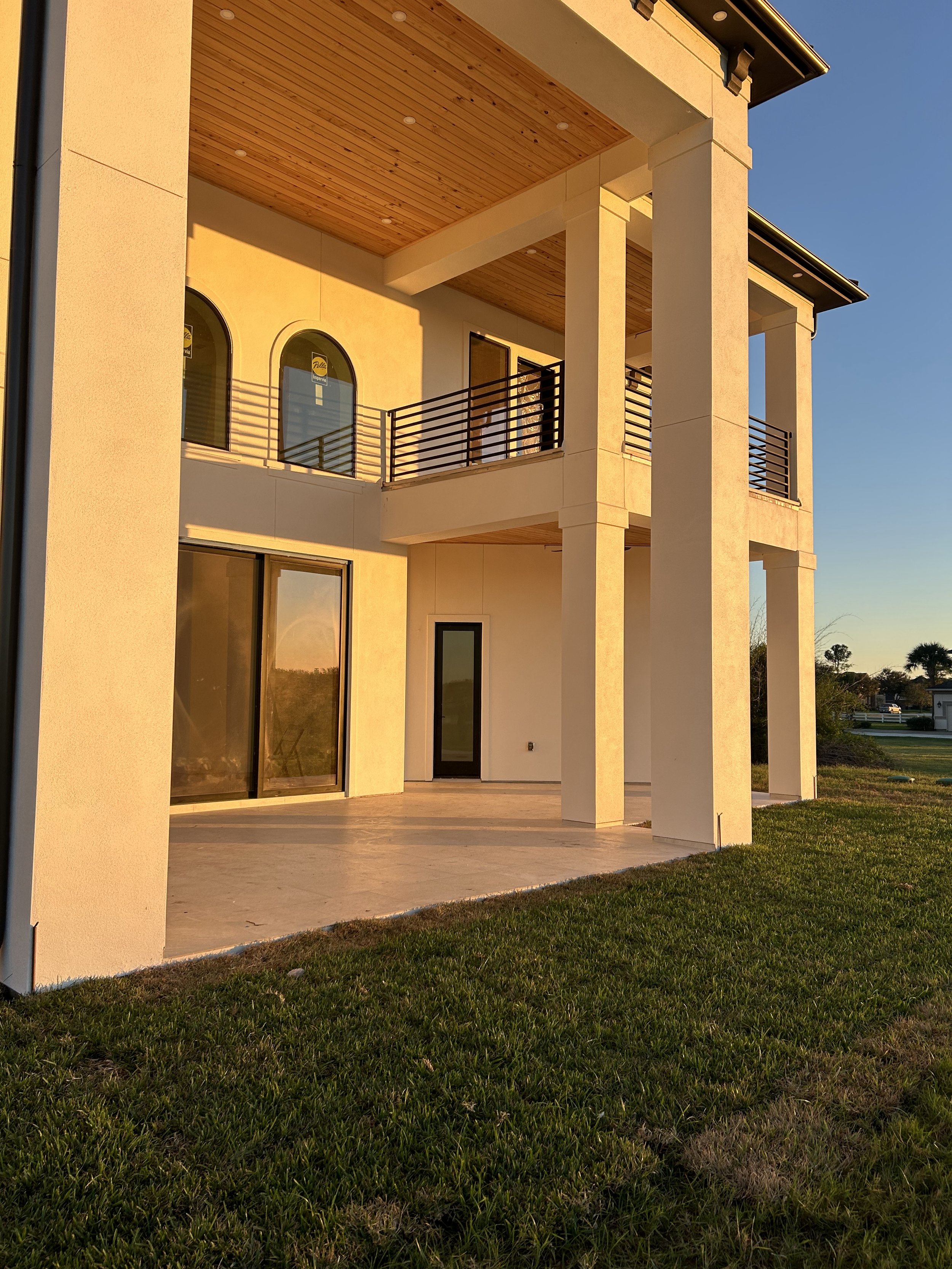 A modern two-story house with a large covered patio, glass sliding doors, and a balcony with black metal railings, illuminated by warm sunlight, with a grassy lawn in the foreground and a clear blue sky.
