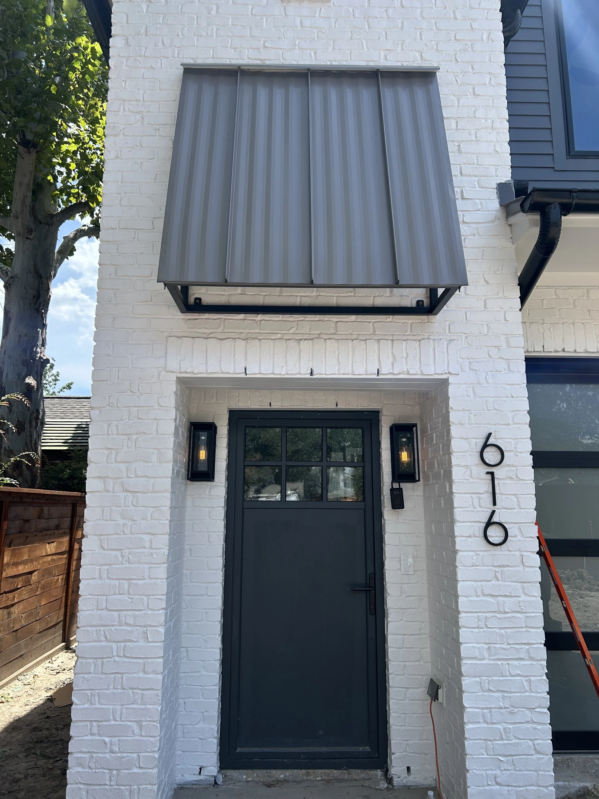 Modern white brick house with a black front door, black wall-mounted lights, metal awning above the door, house number 616 on the right, and a tree on the left side.