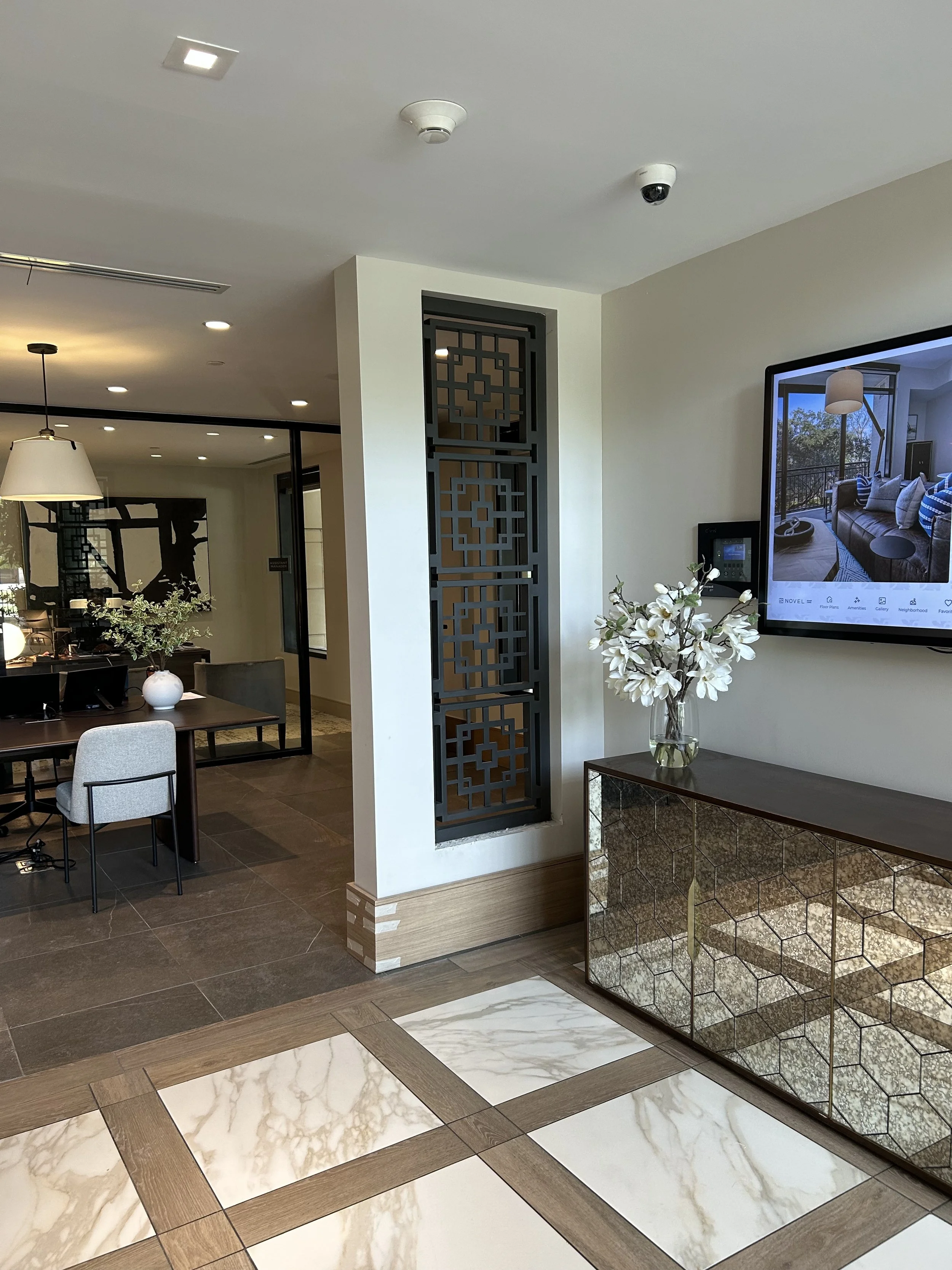 Interior view of a modern living space with a white wall, decorative black metal screen, a flat-screen TV, a vase with white flowers on a mirrored cabinet, and partial views of a dining area with chairs and a table.