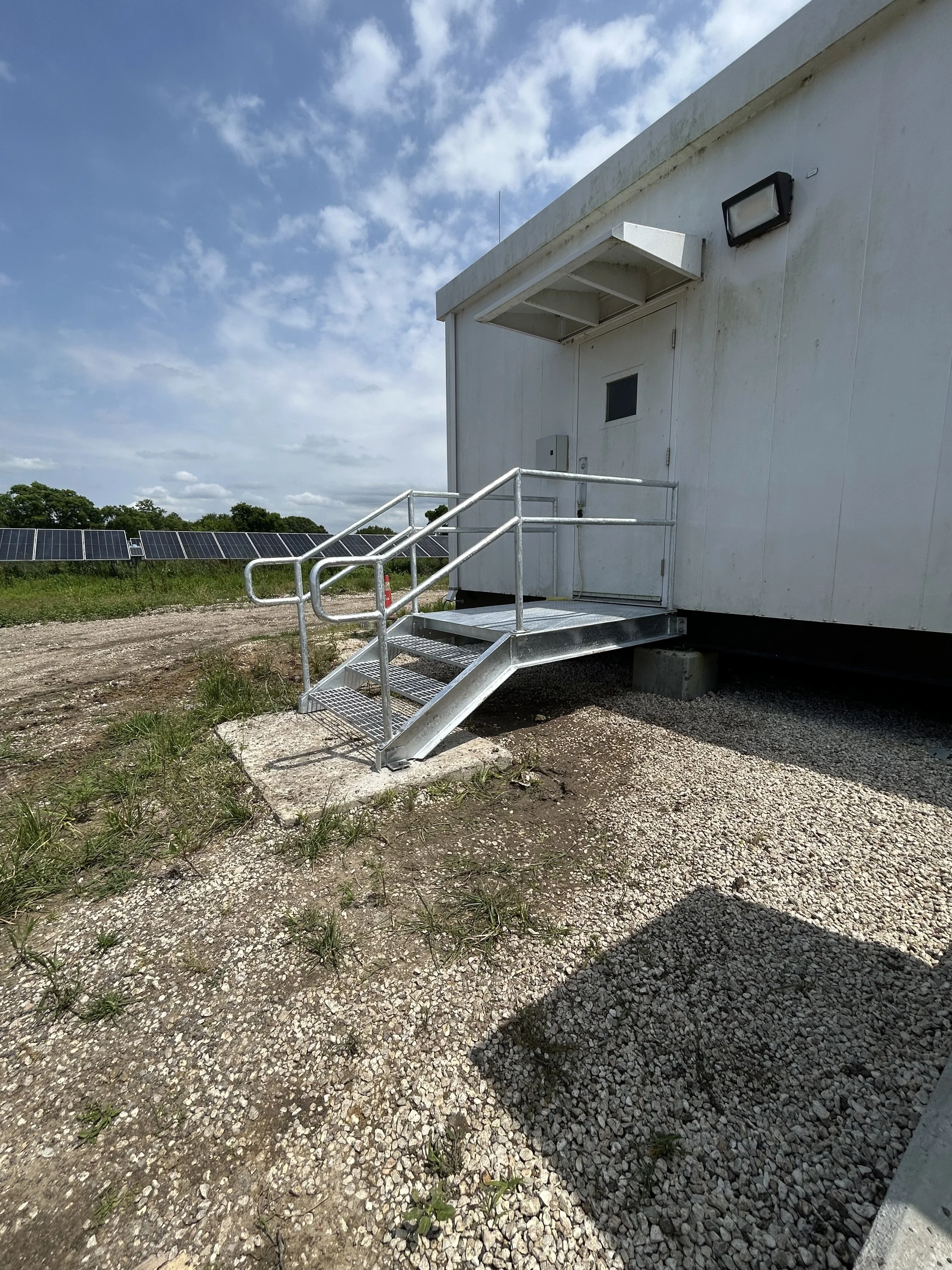 Metal wheelchair ramp leading to a white building with a door and a small window, set outdoors on gravel with solar panels in the background under a partly cloudy sky.