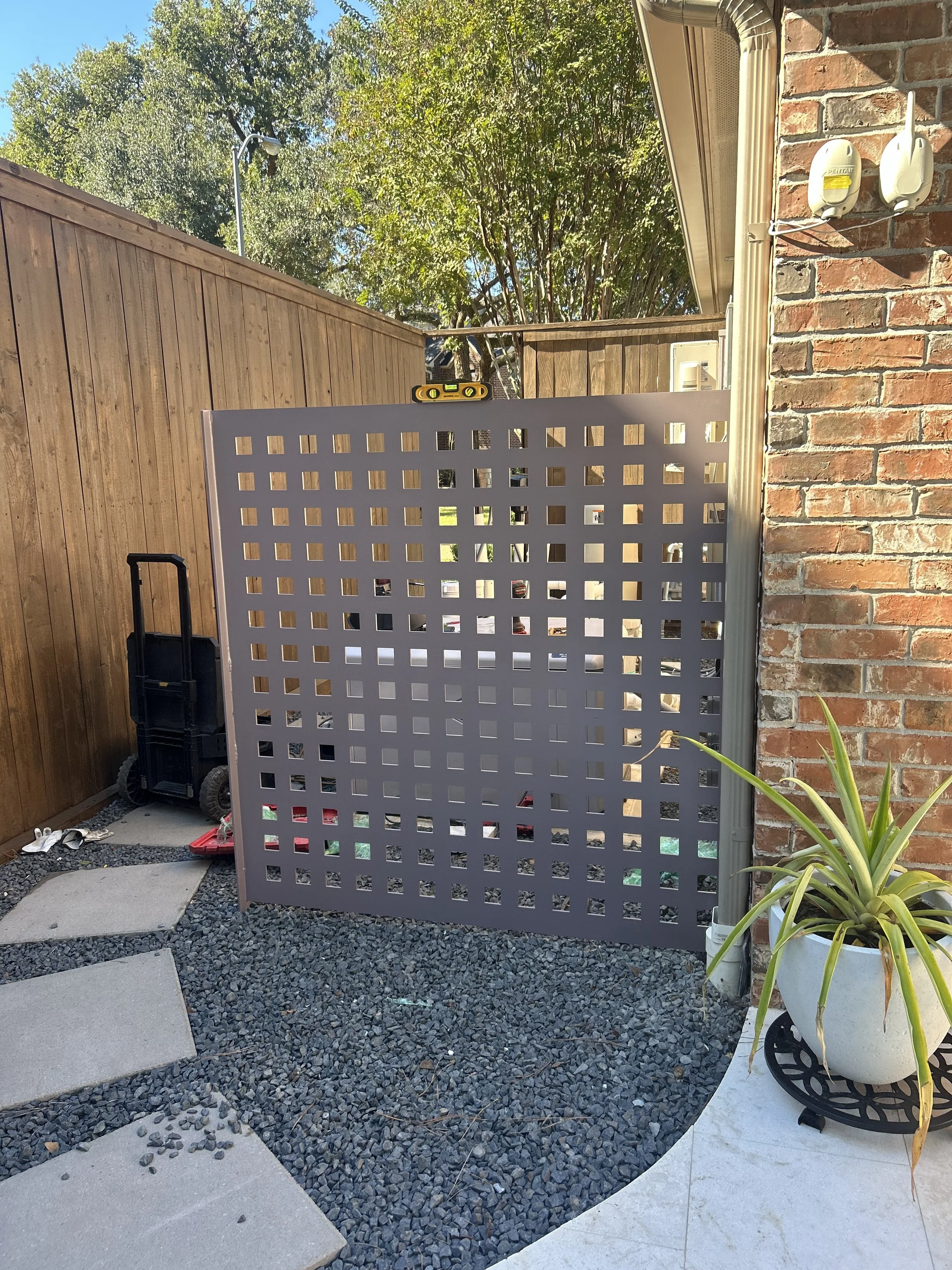 A backyard with a wooden fence, a gray lattice gate, a wheelbarrow, and a potted plant with long green leaves on a black stand. Some tools and a level are on top of the gate. There are concrete pavers on gravel and a concrete patio, with trees in the