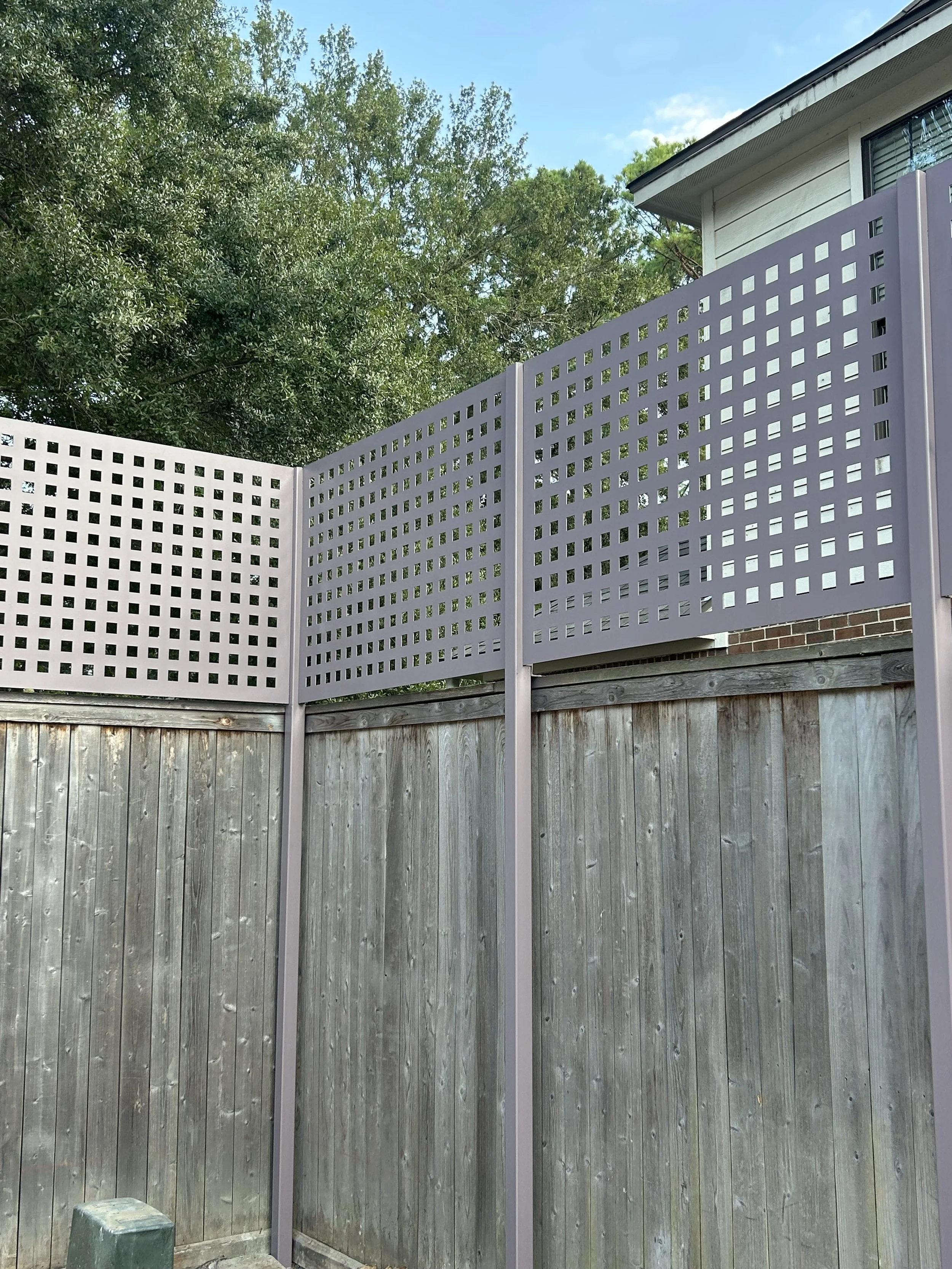 Wooden privacy fence with a purple lattice top, located outdoors near a house and surrounded by green trees.