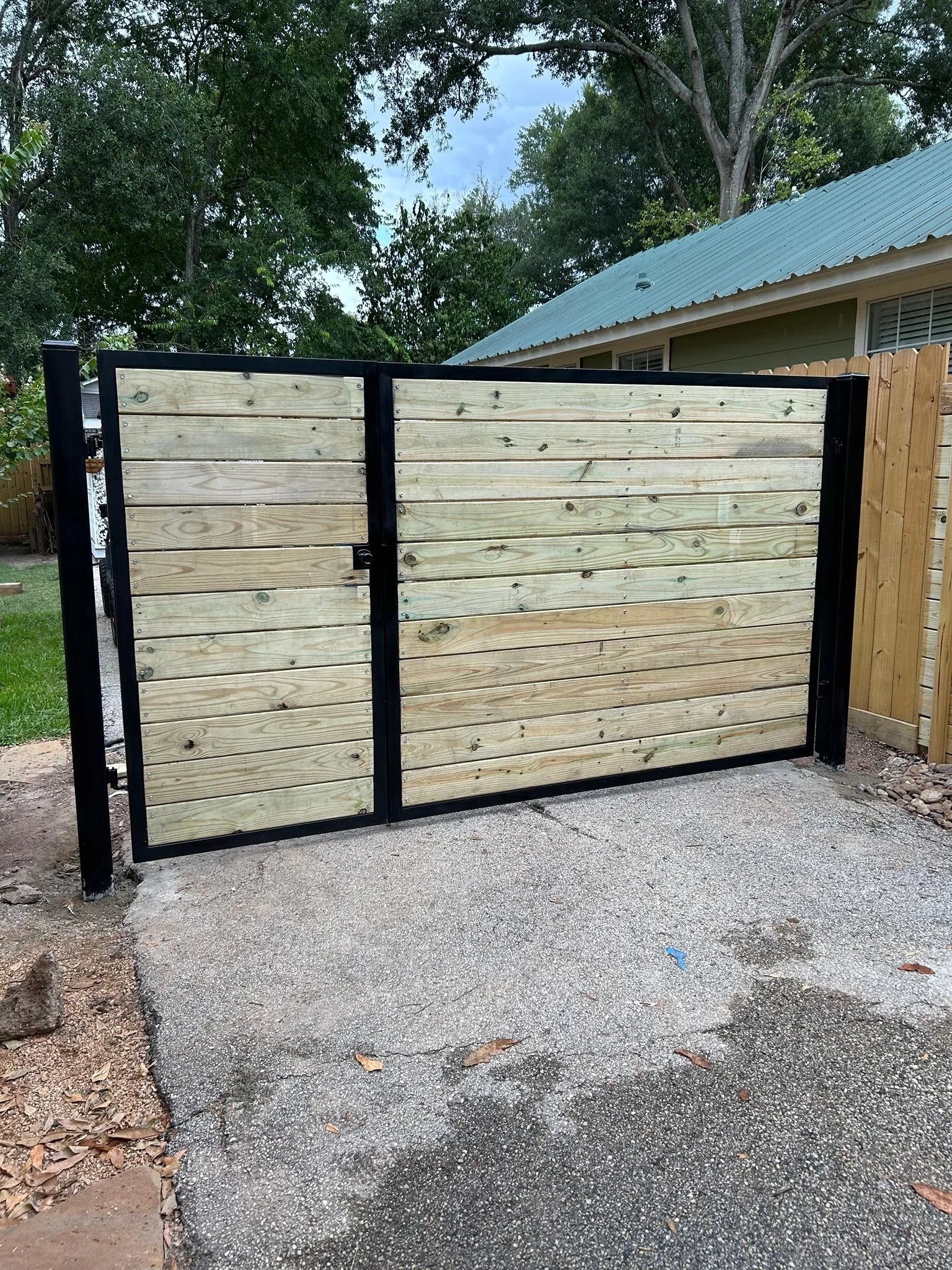 A black metal gate with wooden panels, positioned on a concrete driveway, with a house and trees in the background.