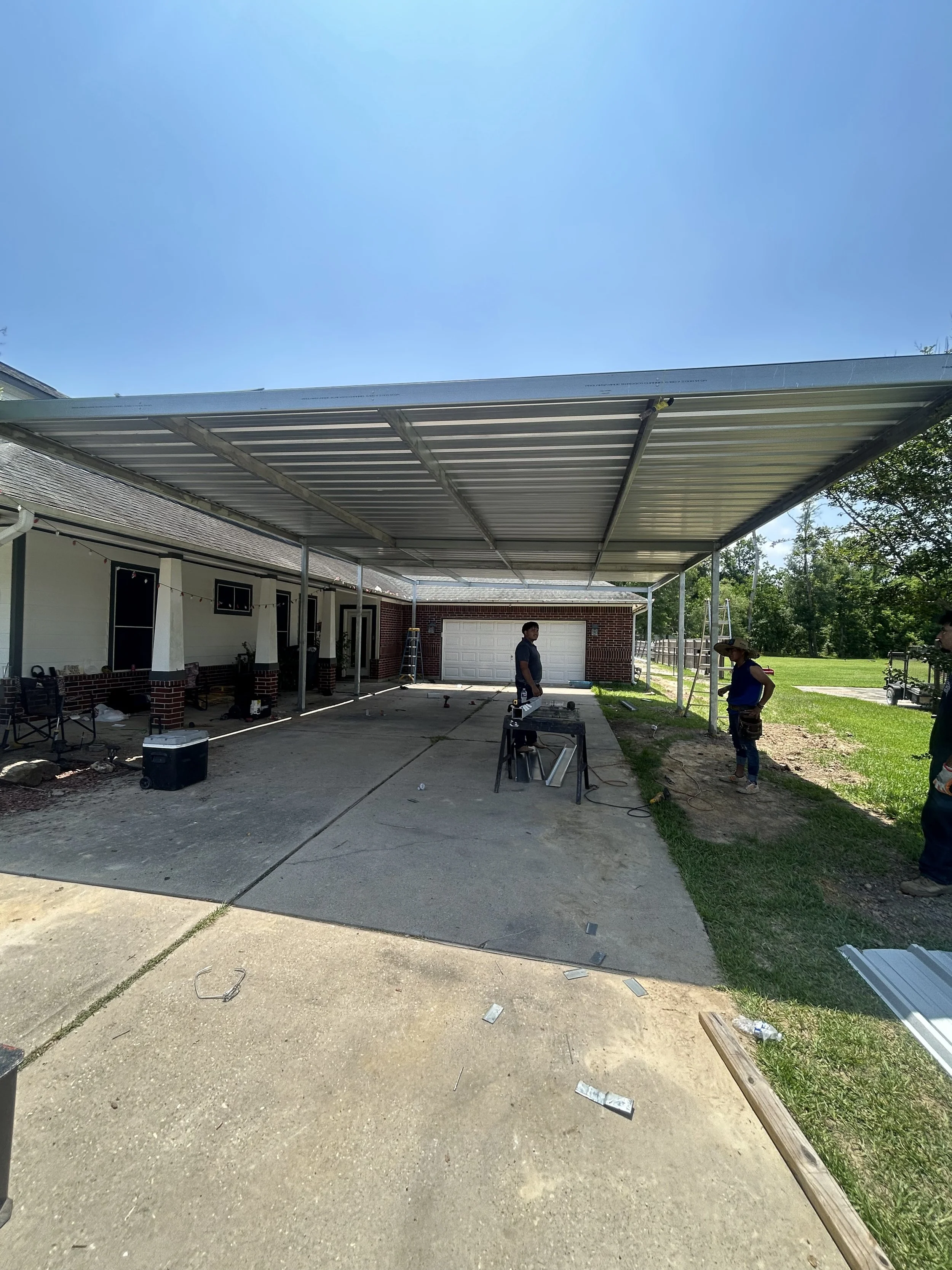 Construction workers installing a metal carport or roof extension in a driveway area of a suburban home, with tools and materials scattered around.