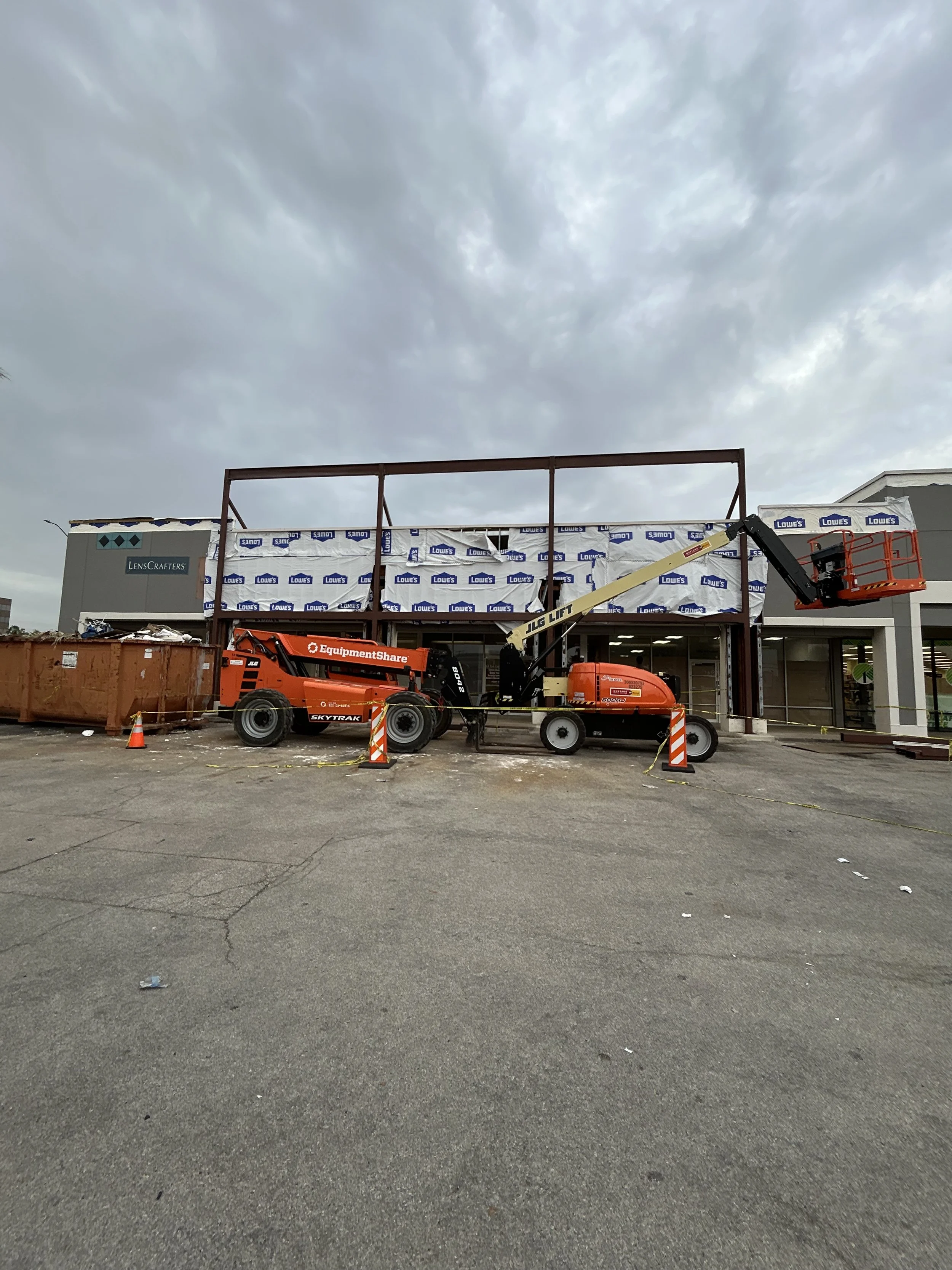 Construction workers are building a storefront at a Lowe’s hardware store, using a lift and equipment, with a sky filled with dark clouds overhead.