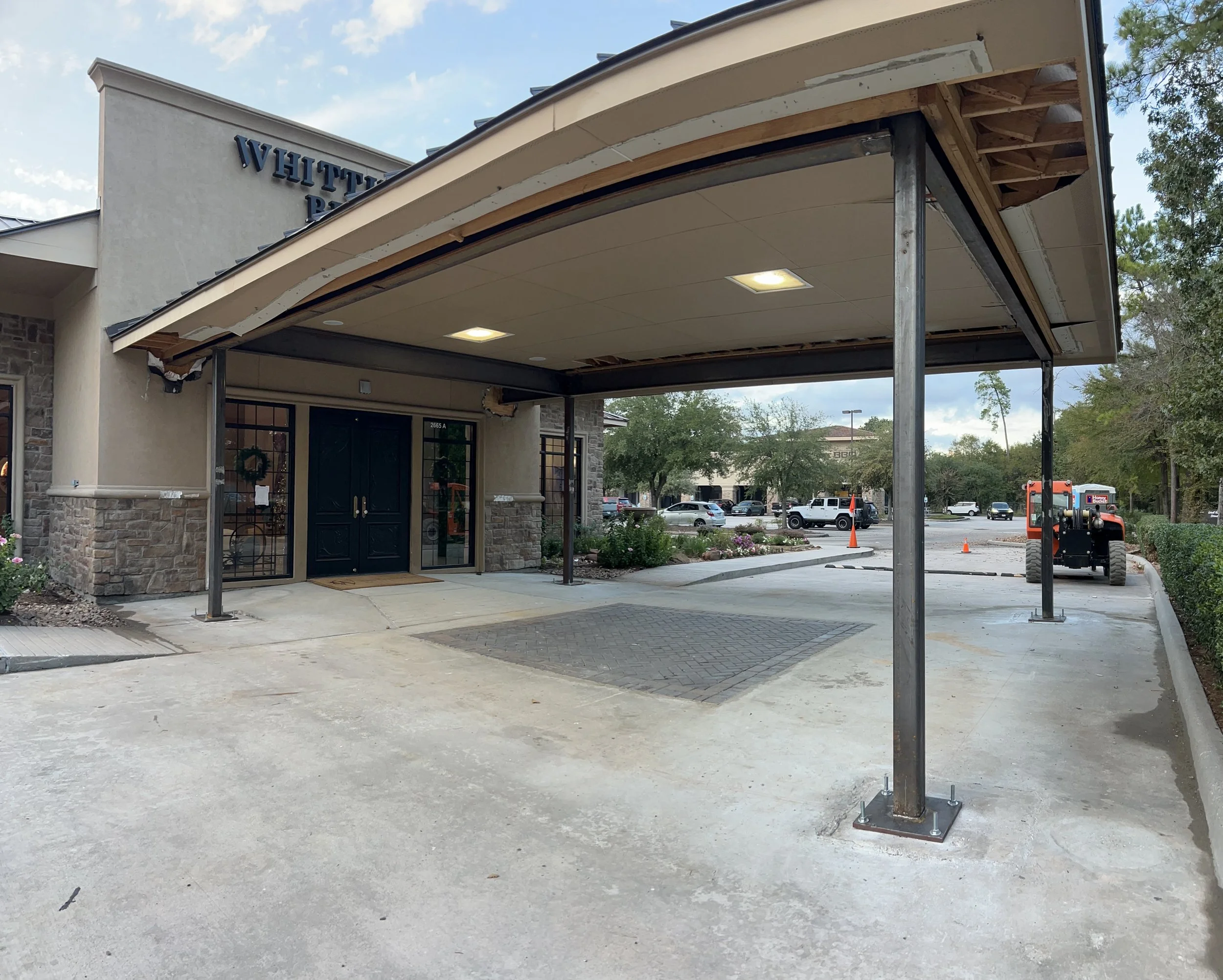 A building with a partially constructed or renovated covered entrance area with metal support beams and a wooden frame supporting the roof. The door is black with glass panels, and the building appears to be a retail or restaurant space. Construction