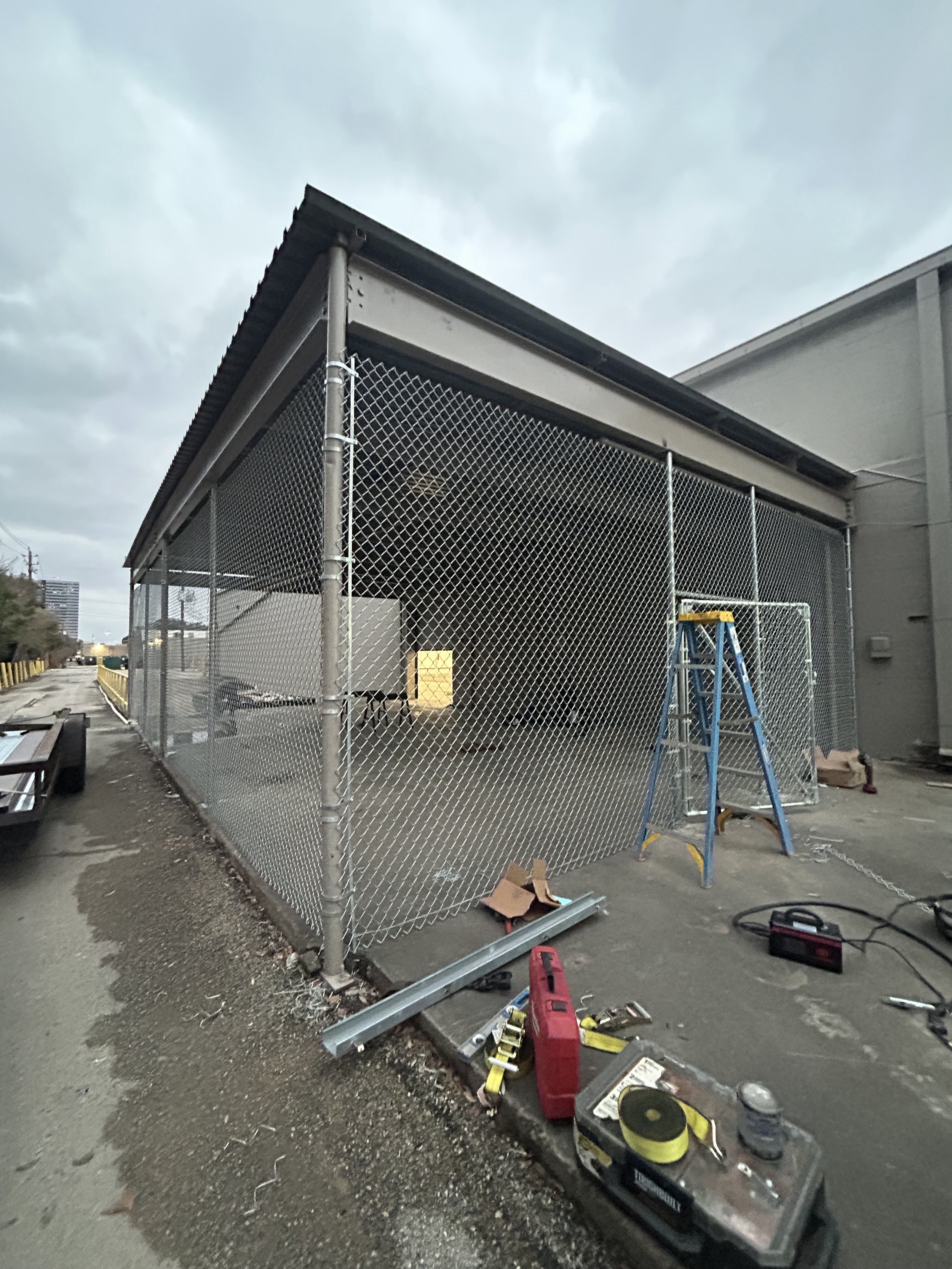 Construction site with a chain-link fence, blue ladder, and tools outside a building under cloudy sky.