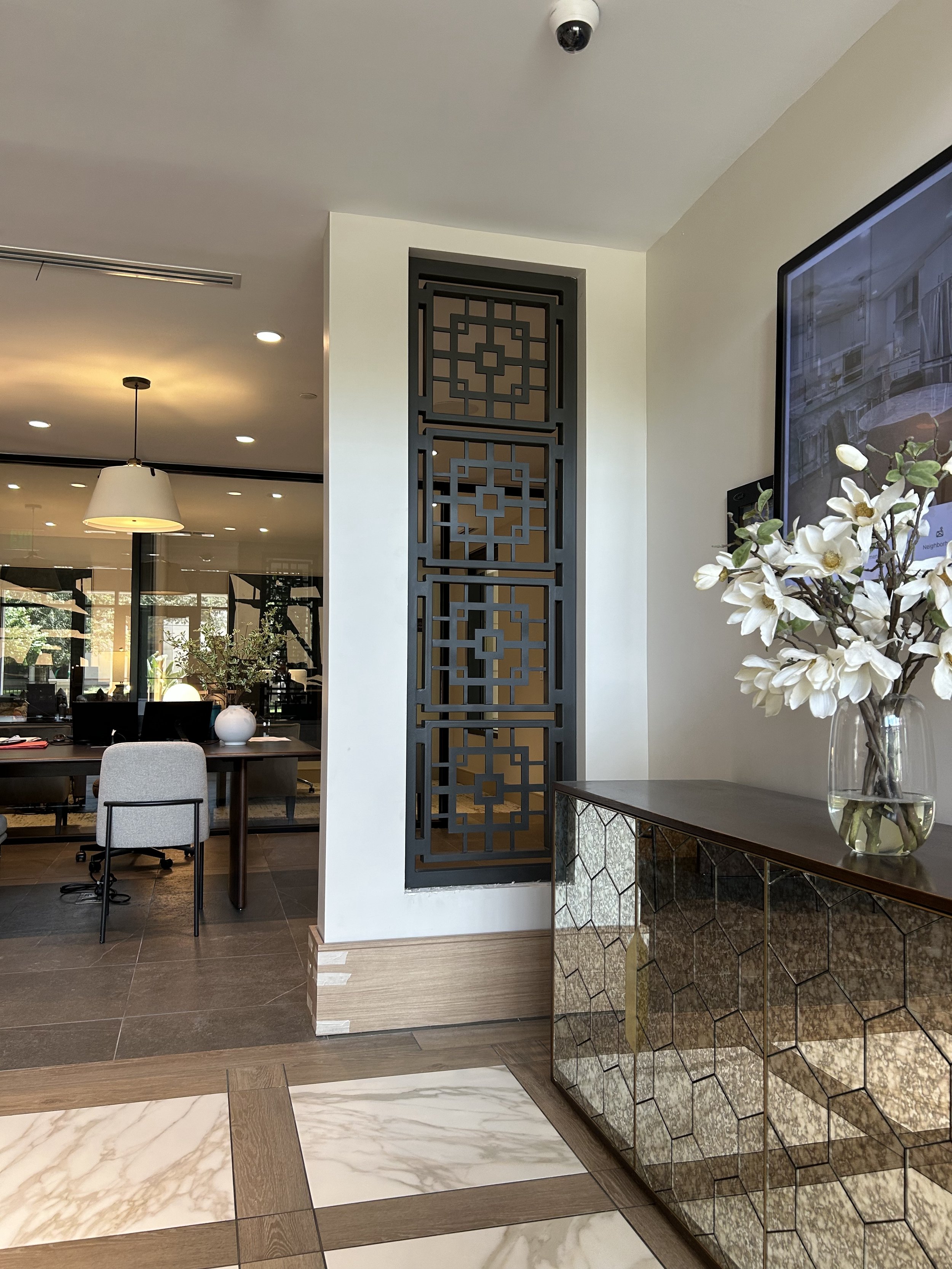 Indoor area with a decorative partition, a cabinet with a geometric pattern, a vase of white flowers, a wall-mounted monitor, and a dining area with chairs and pendant lighting.