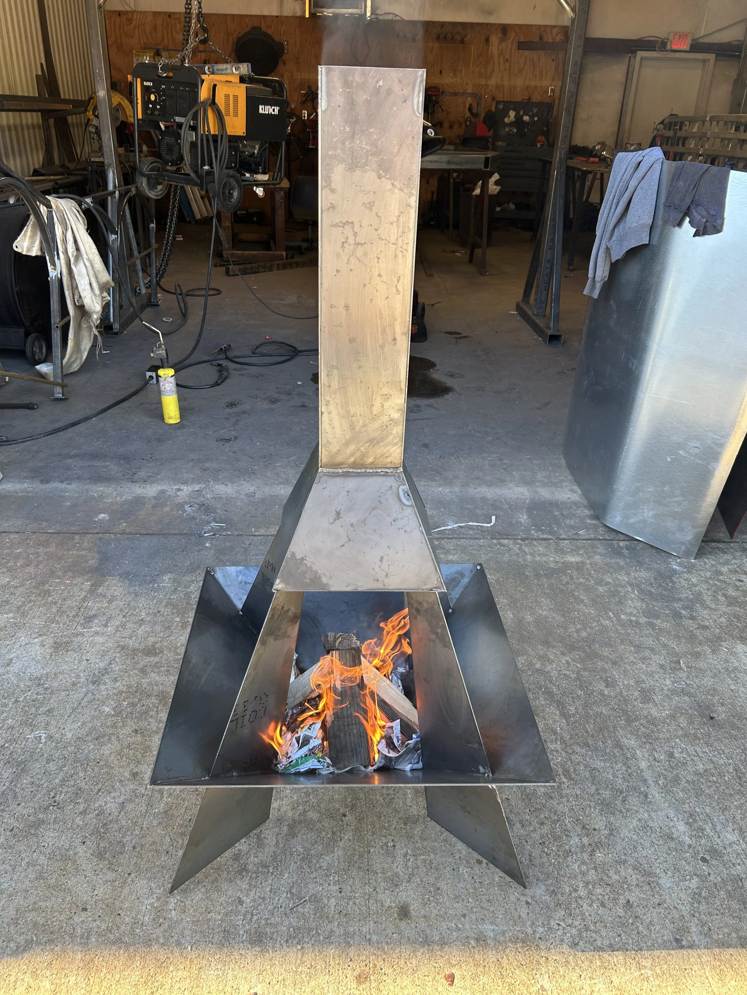 A metal fire pit with an open flame and burning logs, located inside a workshop or garage with tools and equipment in the background.
