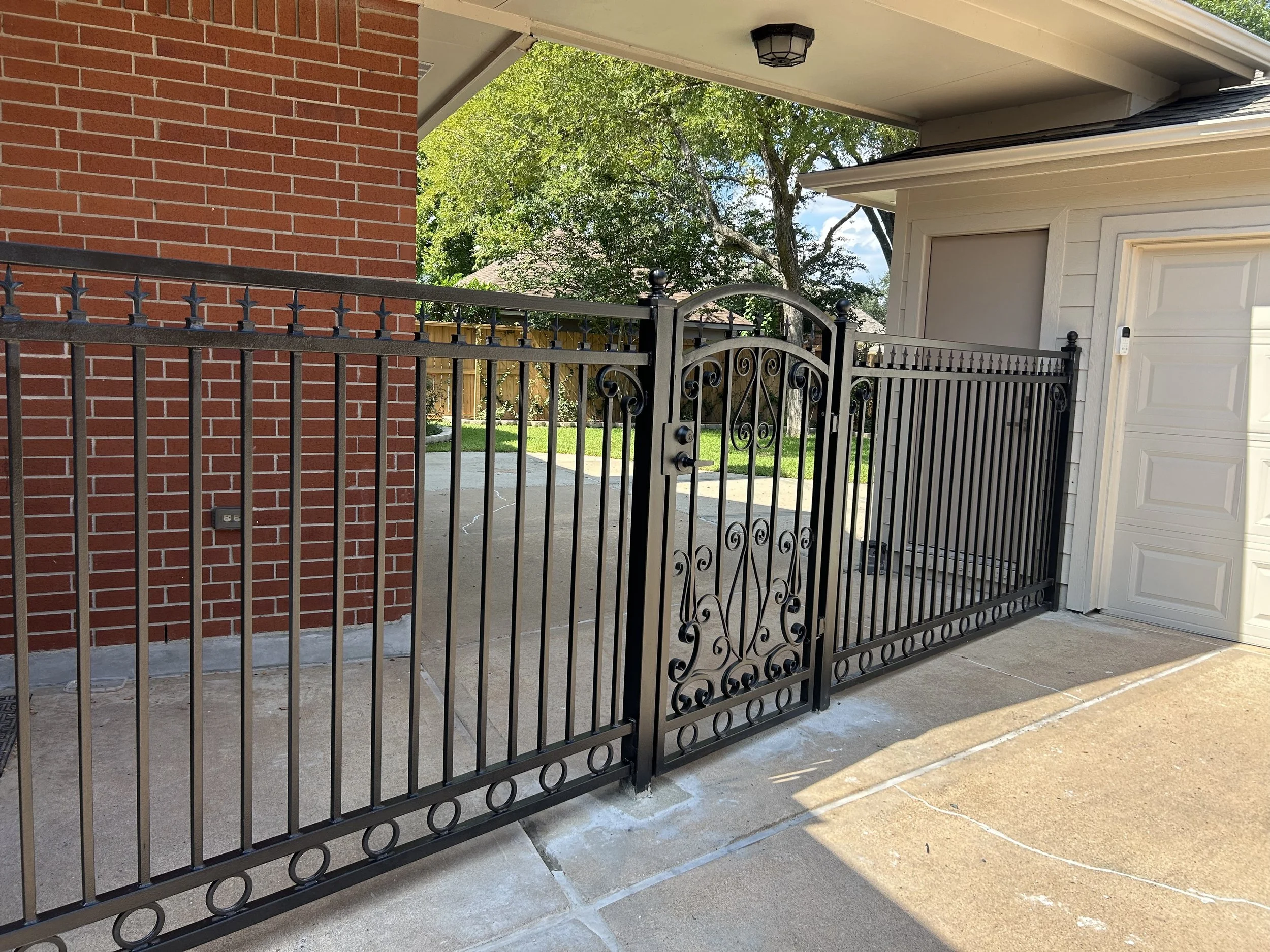 A black metal gate with vertical bars and decorative swirls, located in front of a white garage and brick wall, leading into a backyard with green grass and trees.