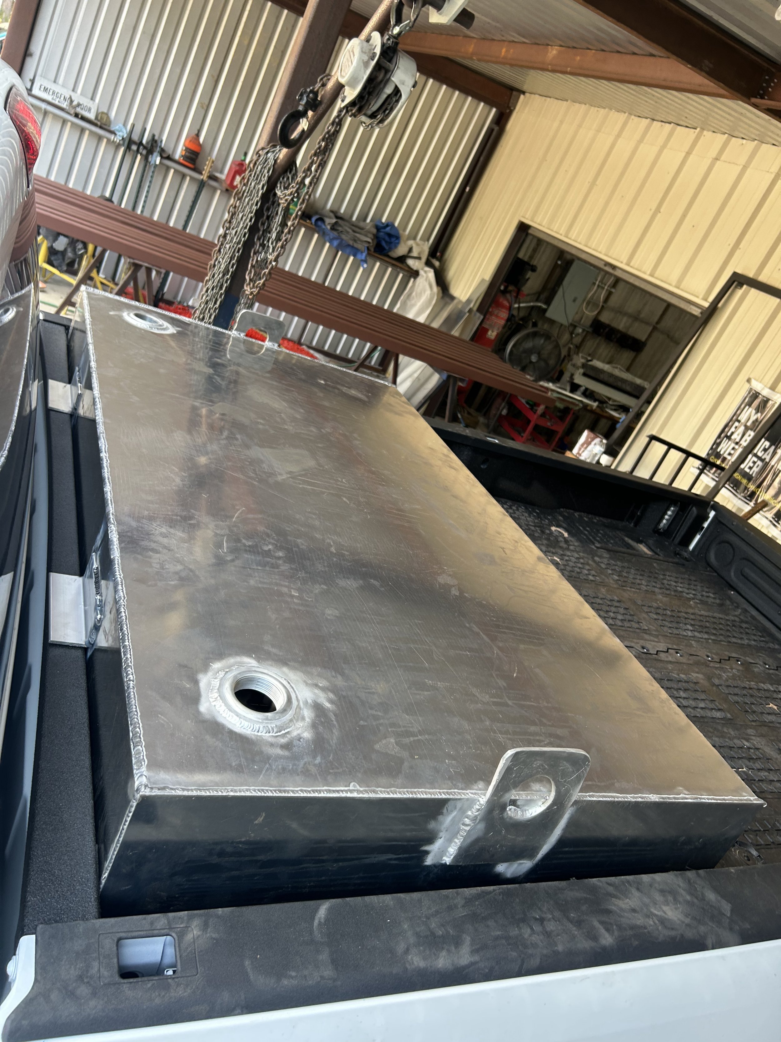 A large rectangular metal tank with a central hole, mounted on a vehicle, with a chain hoist and chains hanging above in a workshop or garage.