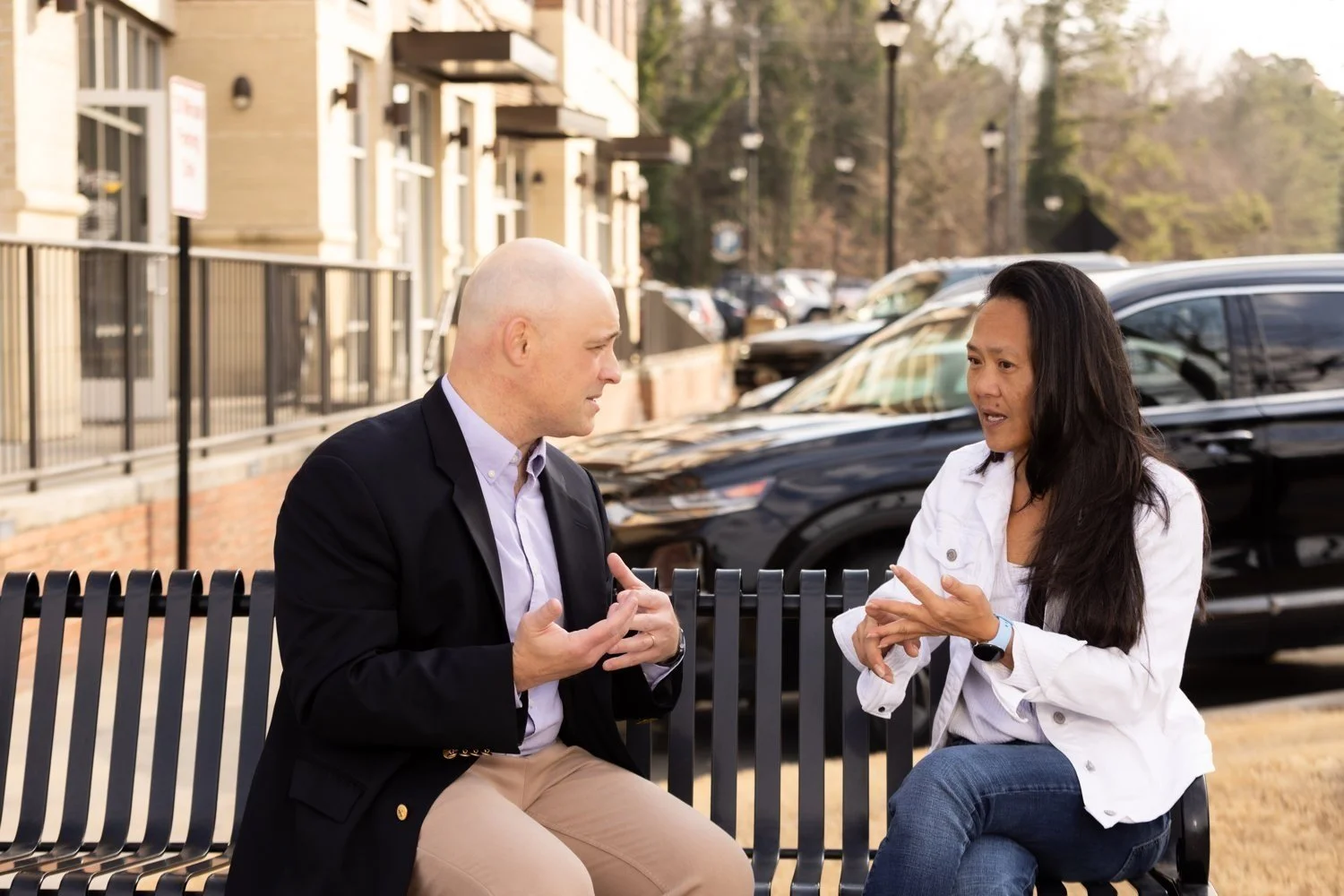 A man and woman sit on a park bench engaged in a conversation. The man is bald, wears a black blazer over a light shirt and beige pants. The woman has long dark hair, wears a white jacket and blue jeans. They are outdoors with parked cars and a residential building in the background.