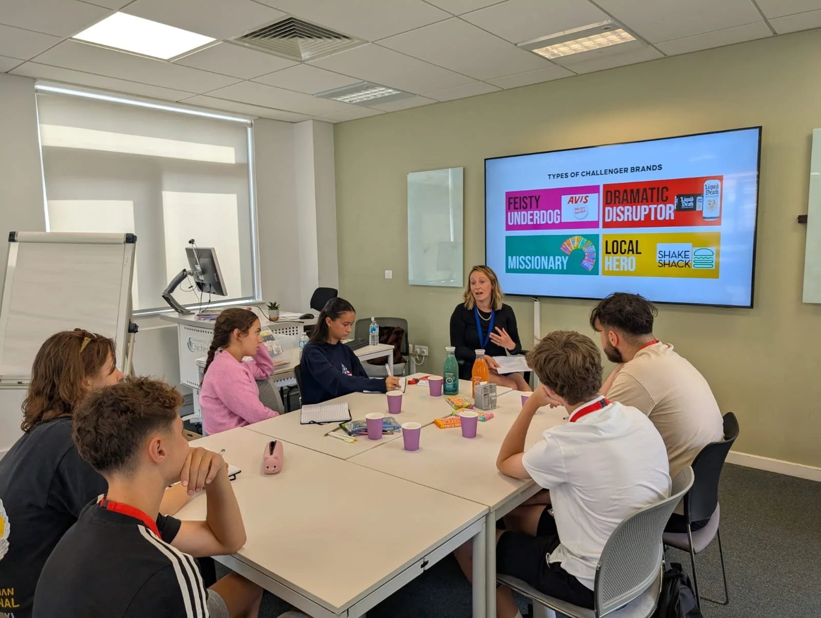 A group of young people attending a meeting or workshop in a conference room, listening to a woman presenting at the front. There is a large screen behind her displaying various brand categories.