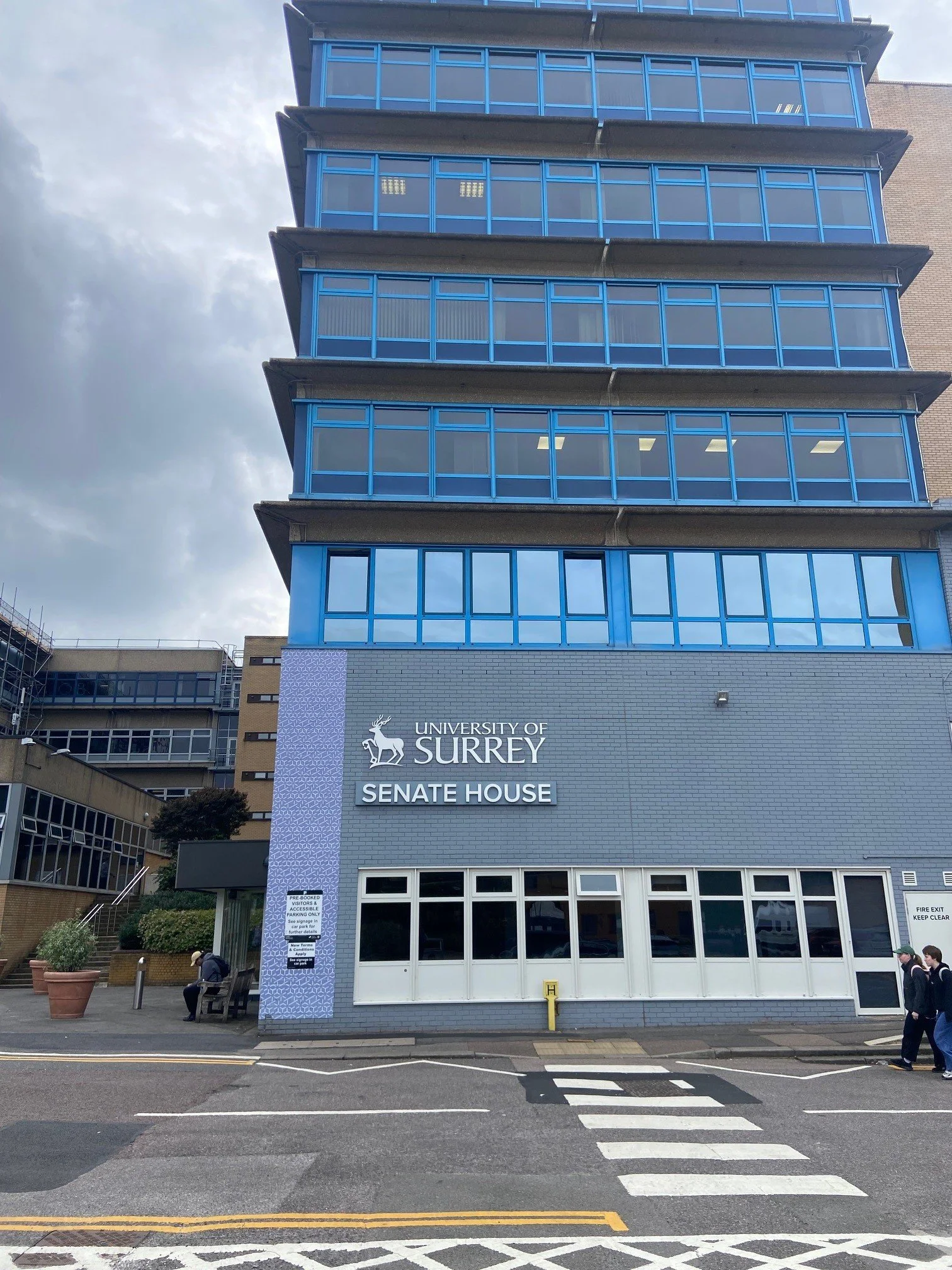 Front of the University of Surrey Senate House building, with a blue glass facade and a gray brick base, and a pedestrian crosswalk in the foreground.