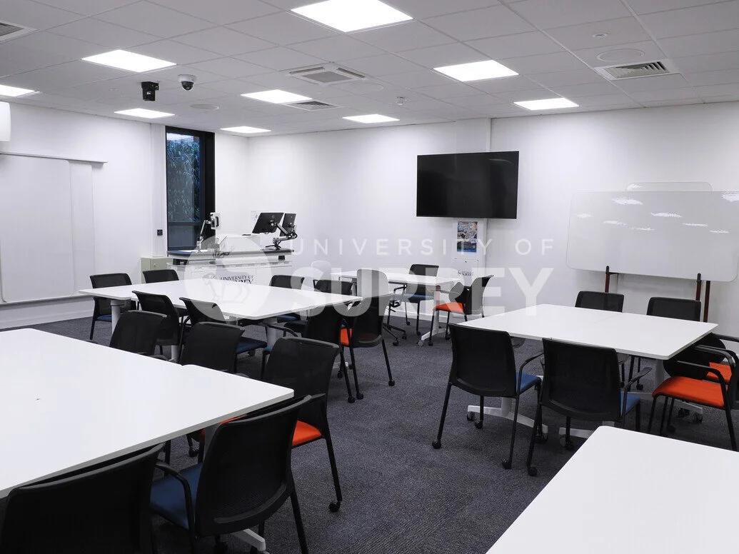 University of surrey conference room with white tables and black chairs, some with orange cushions. Features a large wall-mounted television, whiteboard, two desktop computers on a desk near a window, and a small stand with posters or papers.
