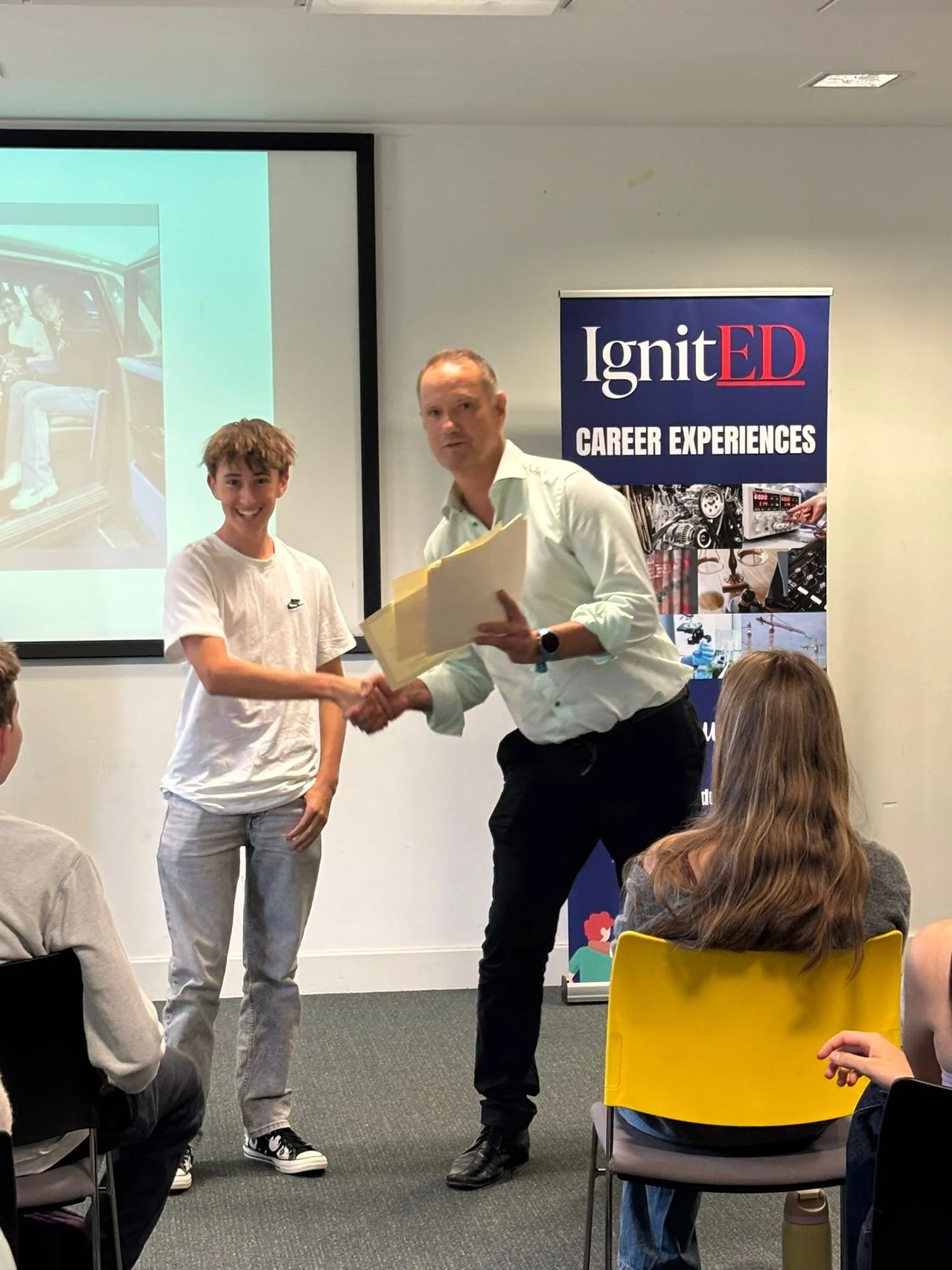 A boy receiving an award and shaking hands with a man during a presentation or ceremony in a classroom or conference room setting. There are seated audience members and a banner with the text 'IgnitED Career Experiences' in the background.