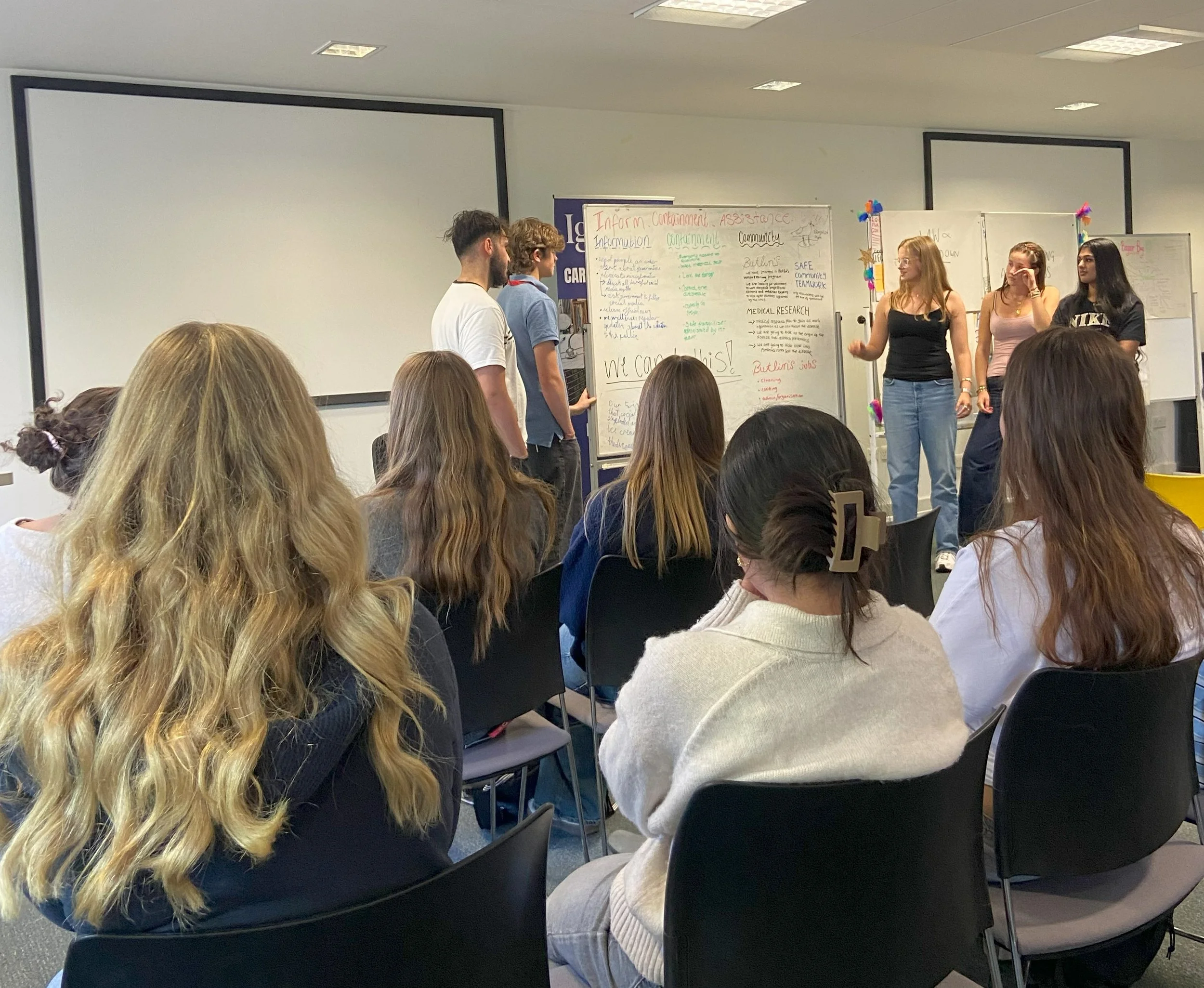Group of students presenting a project to an audience in a classroom. Four students are standing at the front near a large whiteboard with colorful notes. The audience is seated and attentively listening.