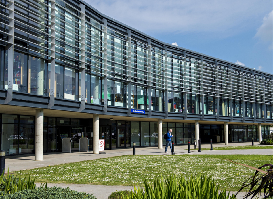 University of Brighton Modern university or office building with glass facade, a person walking on sidewalk in front, and well-maintained landscaping with green grass and bushes.