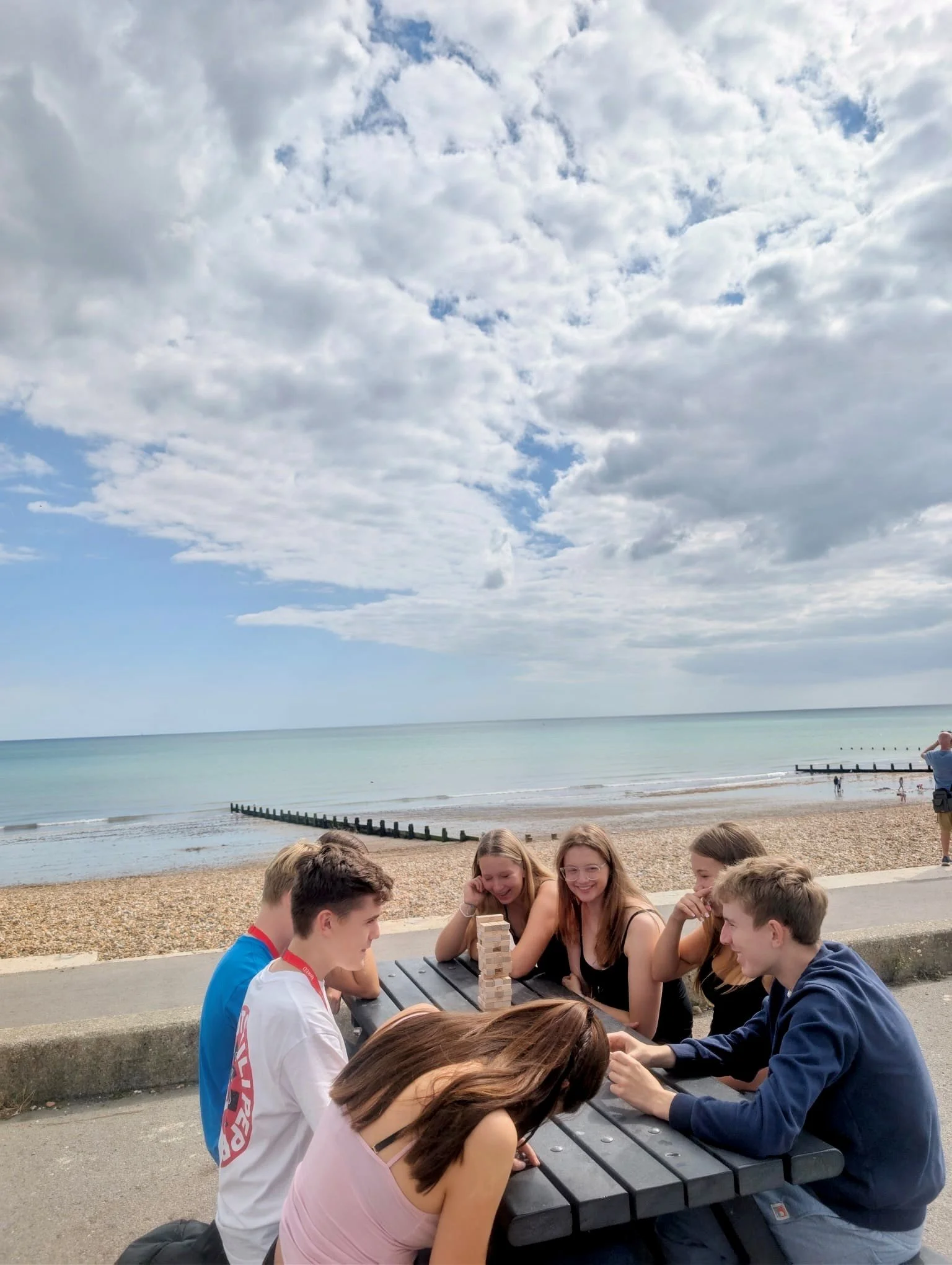 A group of young friends playing Jenga at a picnic table near a beach, with the ocean, sky, clouds, and breakwaters in the background.