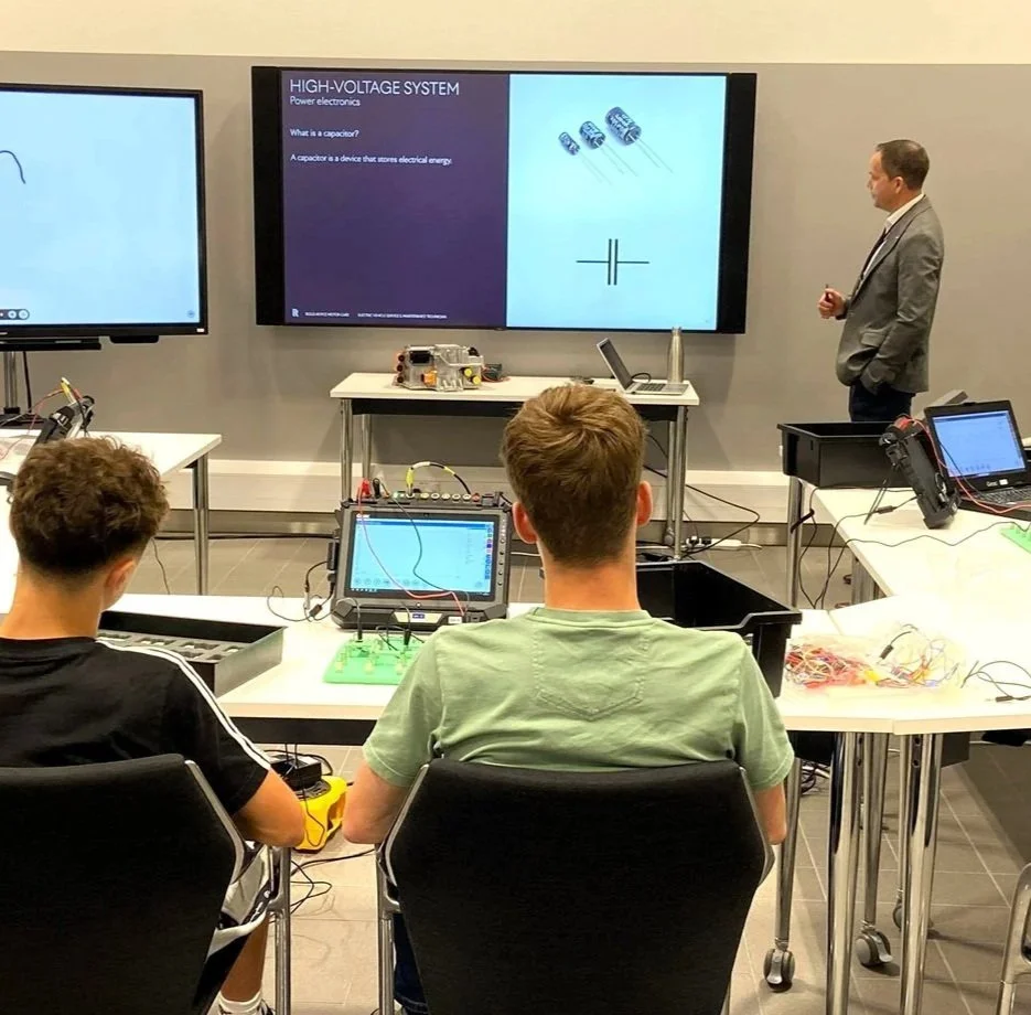 A classroom or lab with students learning about high-voltage systems and capacitors, with a teacher giving a presentation on the electronic components displayed on a screen.