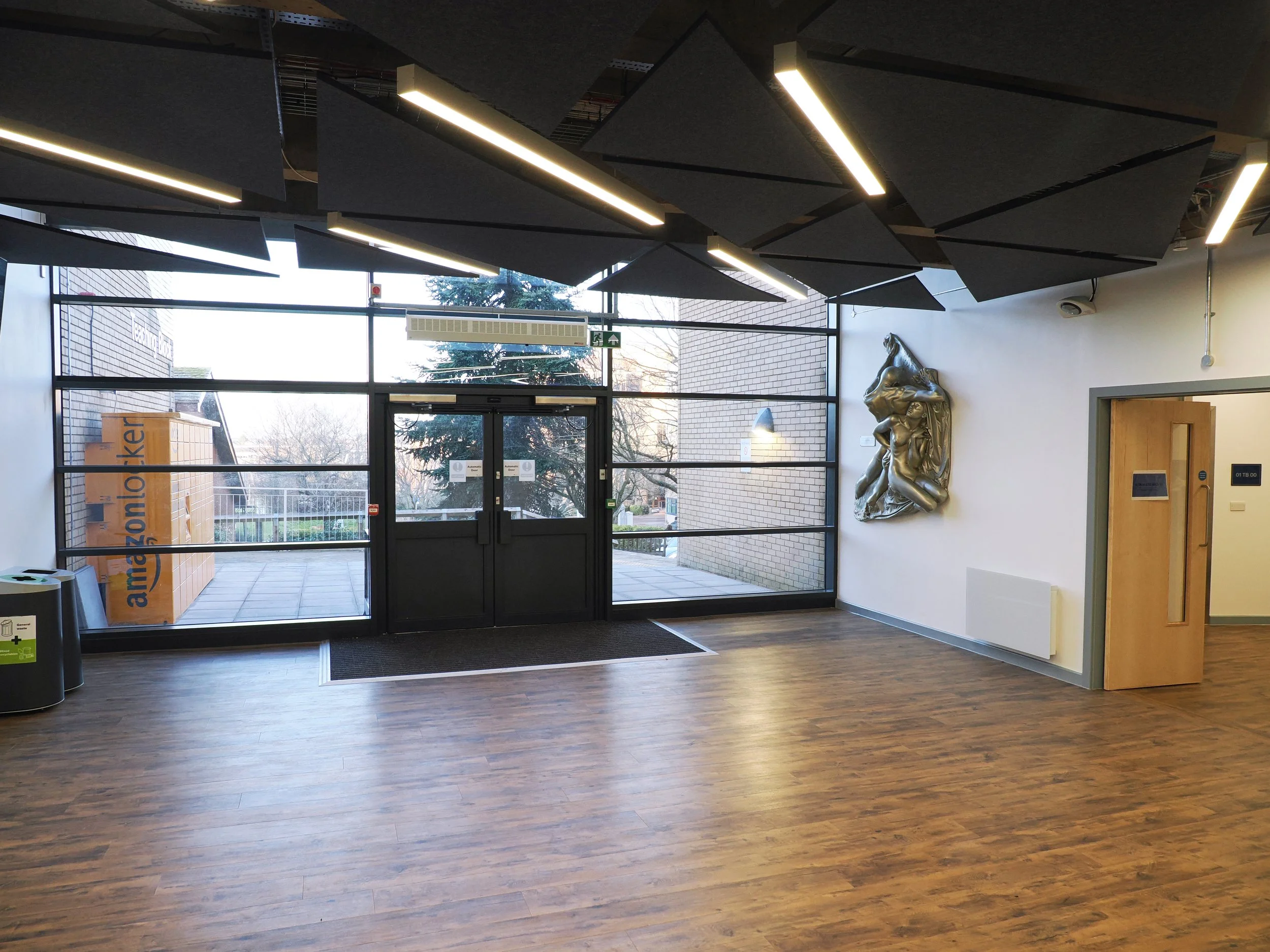 Interior view of a modern office building lobby with wooden flooring, large glass entrance doors, decorative ceiling panels with integrated lighting, a metallic wall sculpture, and an Amazon Locker outside visible through the glass.