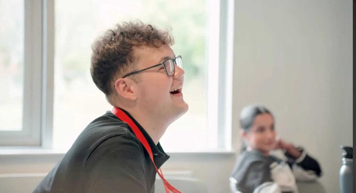 A young man with curly hair and glasses smiling and laughing, sitting at a table in a bright room with a large window behind him, with a woman in the background.
