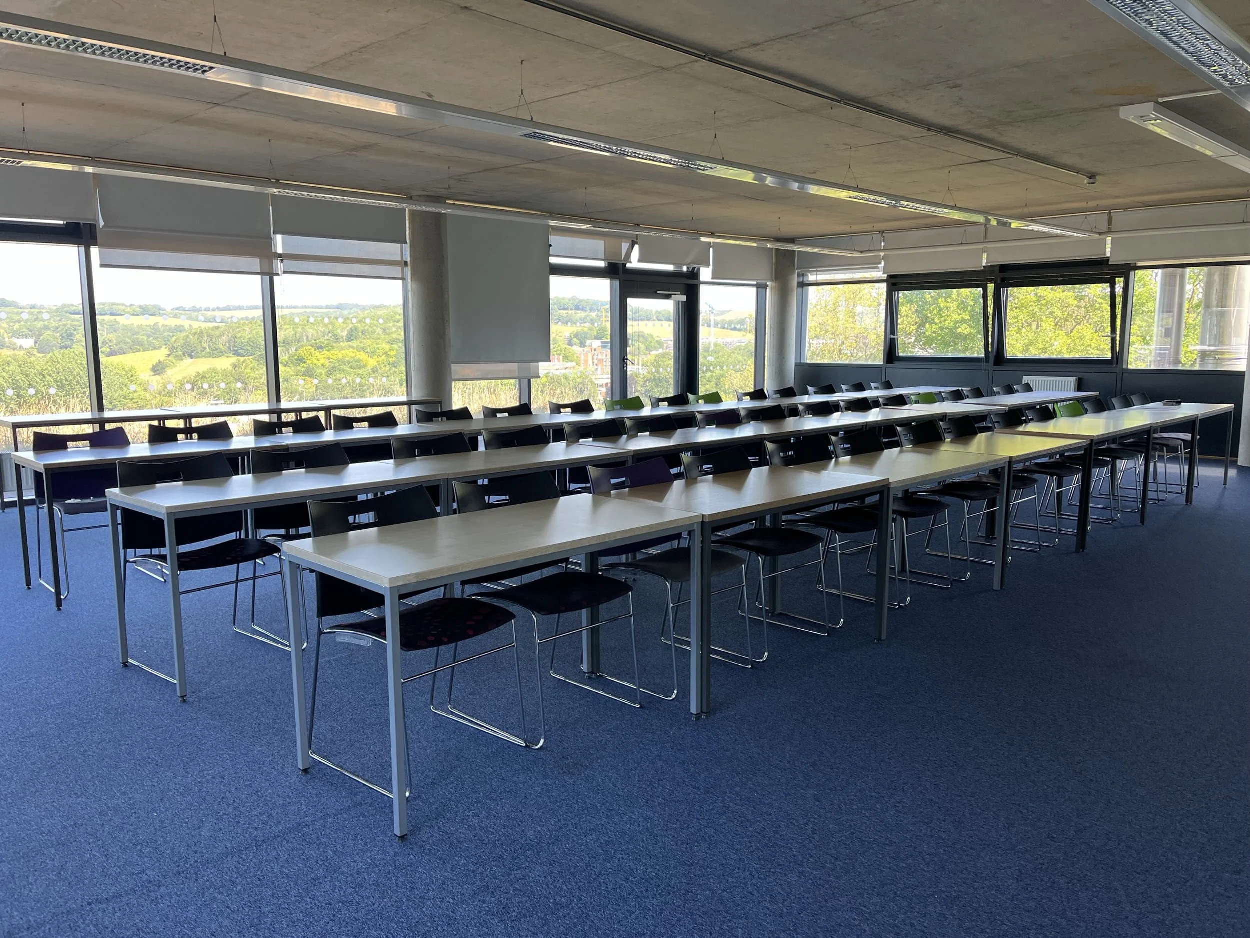 University of Brighton conference room with white tables arranged in a U-shape, black chairs, large windows showing a green landscape, blue carpet, and ceiling lights.