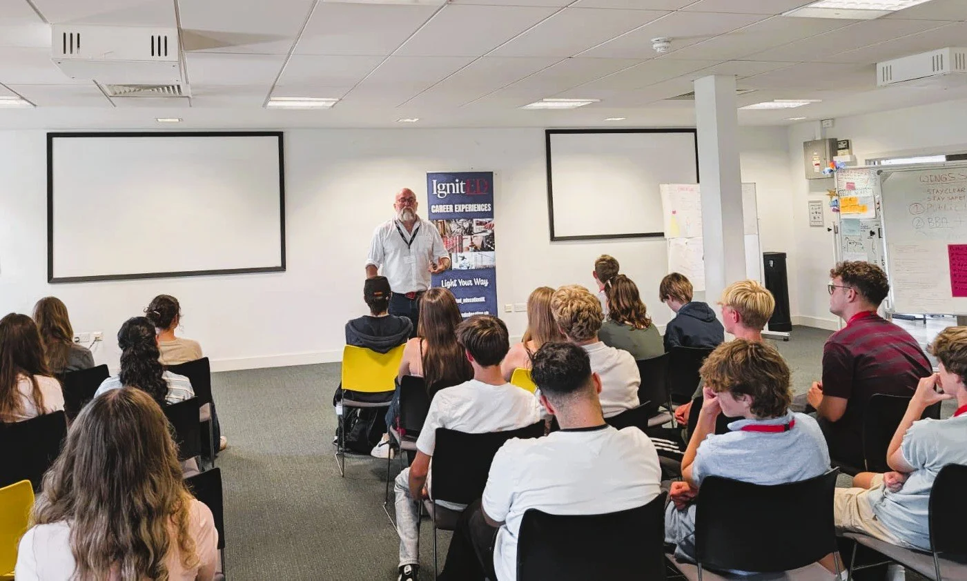 A classroom with students seated facing a male instructor standing near a whiteboard and a banner. There are two large empty screens on the wall behind the instructor.