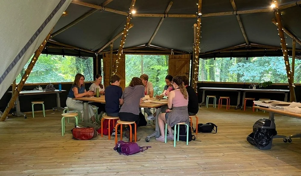 Group of people sitting around a table inside a tents in a forested area, with string lights hanging from the ceiling.
