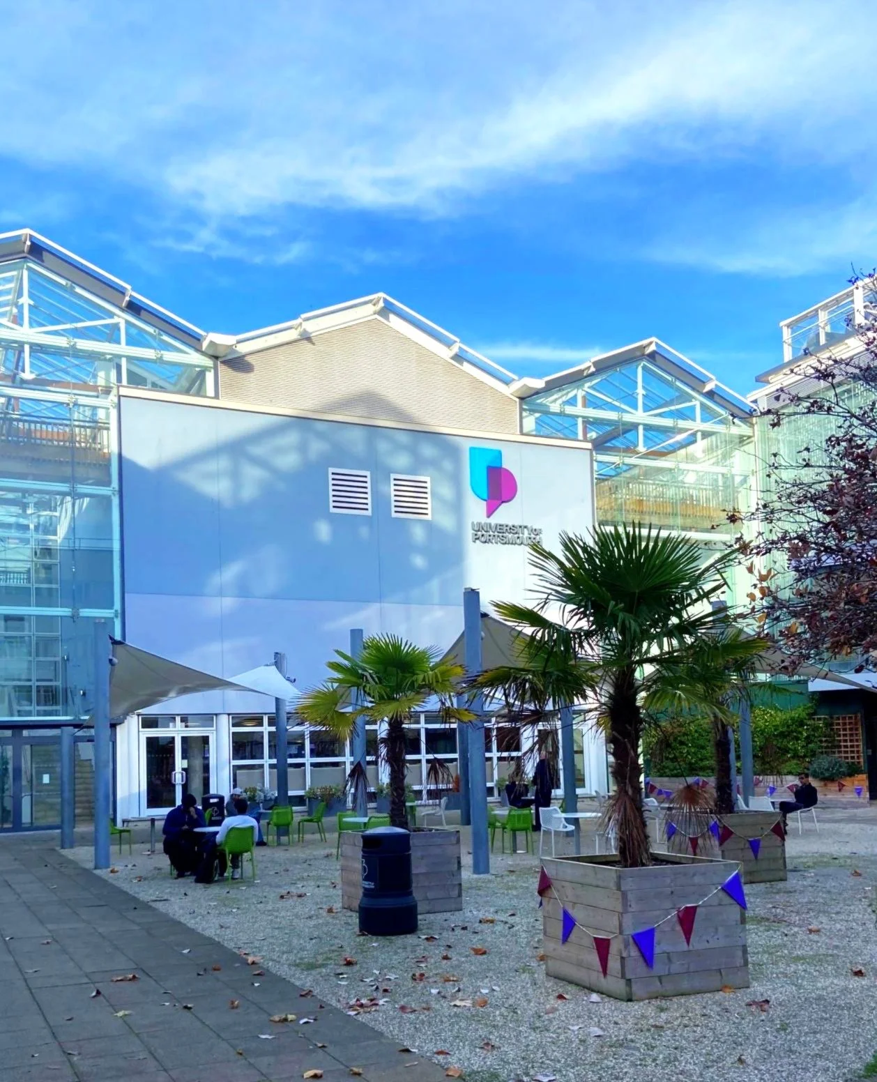 Outdoor scene at University of Portsmouth showing people sitting at green chairs under white canopies, palm trees in wooden planters decorated with red, white, and blue bunting, and a modern building with glass and metal architecture under a blue sky