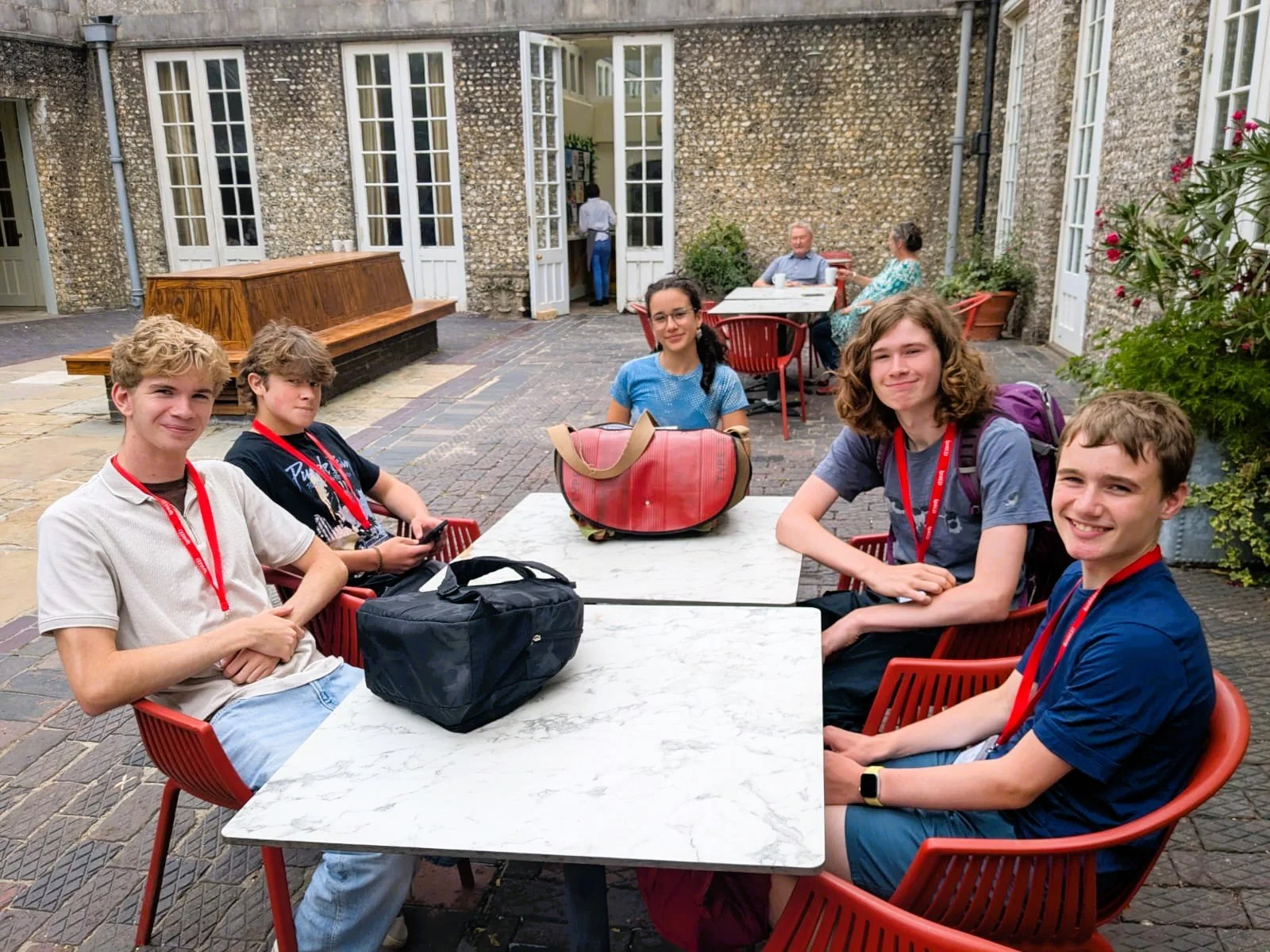Group of six teenagers sitting around outdoor table, with two adults seated in the background at another table, in a courtyard with stone walls and open doors.