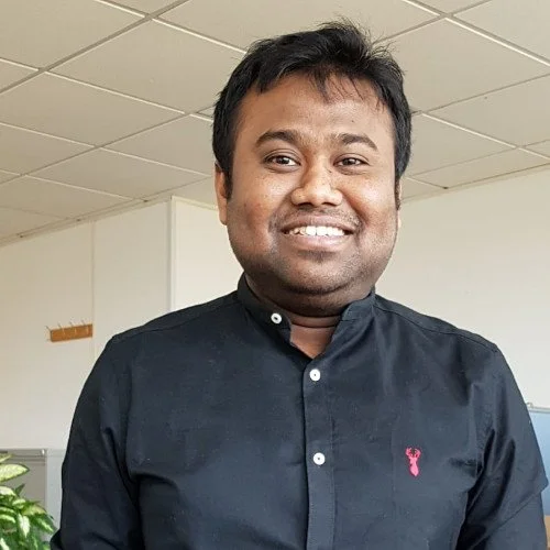 A man with dark hair and brown skin smiling, wearing a black button-up shirt in an indoor setting.