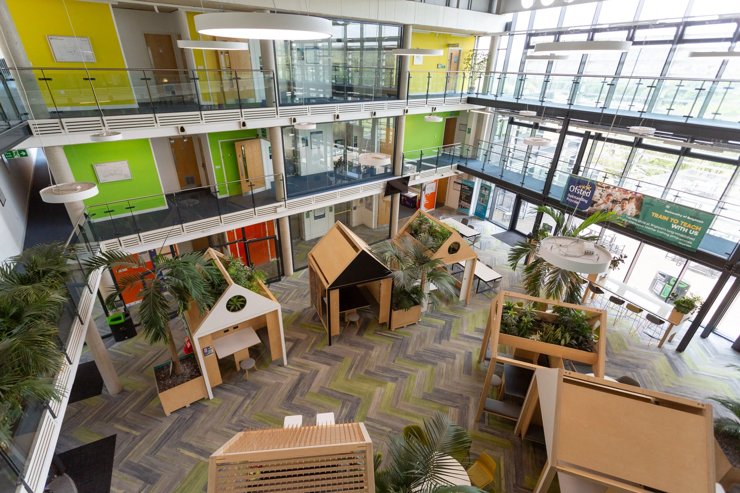 University of Brighton of a modern, colorful educational space with multiple levels, small wooden shelters with plants, large windows, and green and yellow walls.