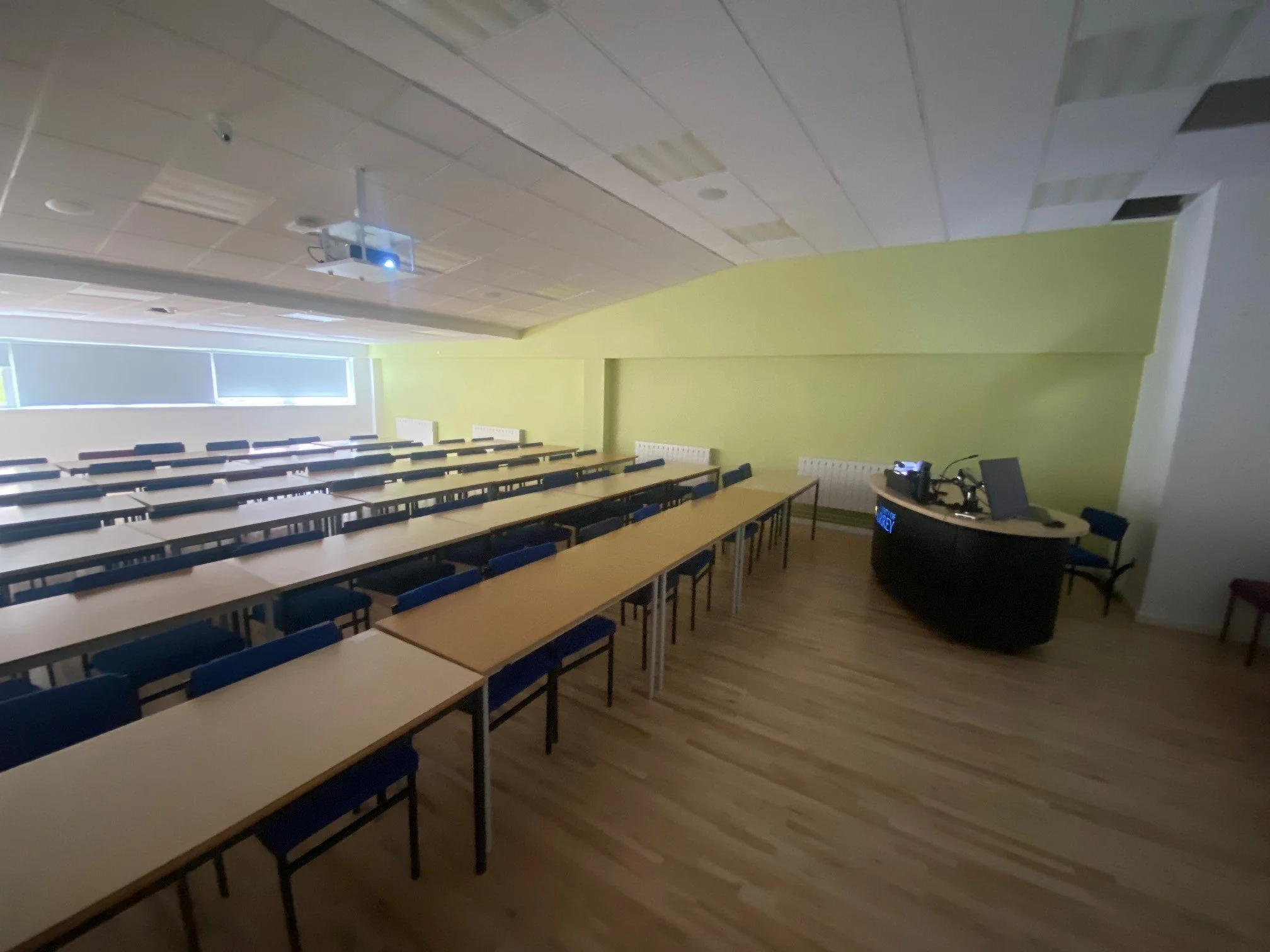 university of surrey classroom with multiple rows of desks and chairs, a green wall, a curved teacher's desk with a computer and projector, and windows with blinds at the back.