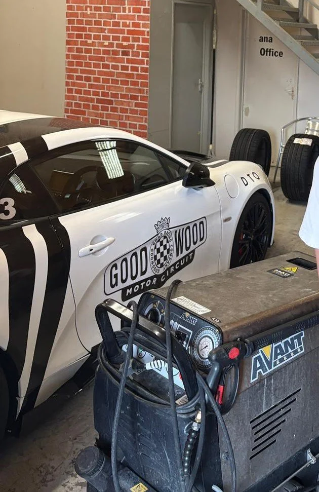 A race car with 'Goodwood Motor Circuit' logo on the side. The car is white with black stripes and has number 3. It is inside a garage with tires and equipment around.