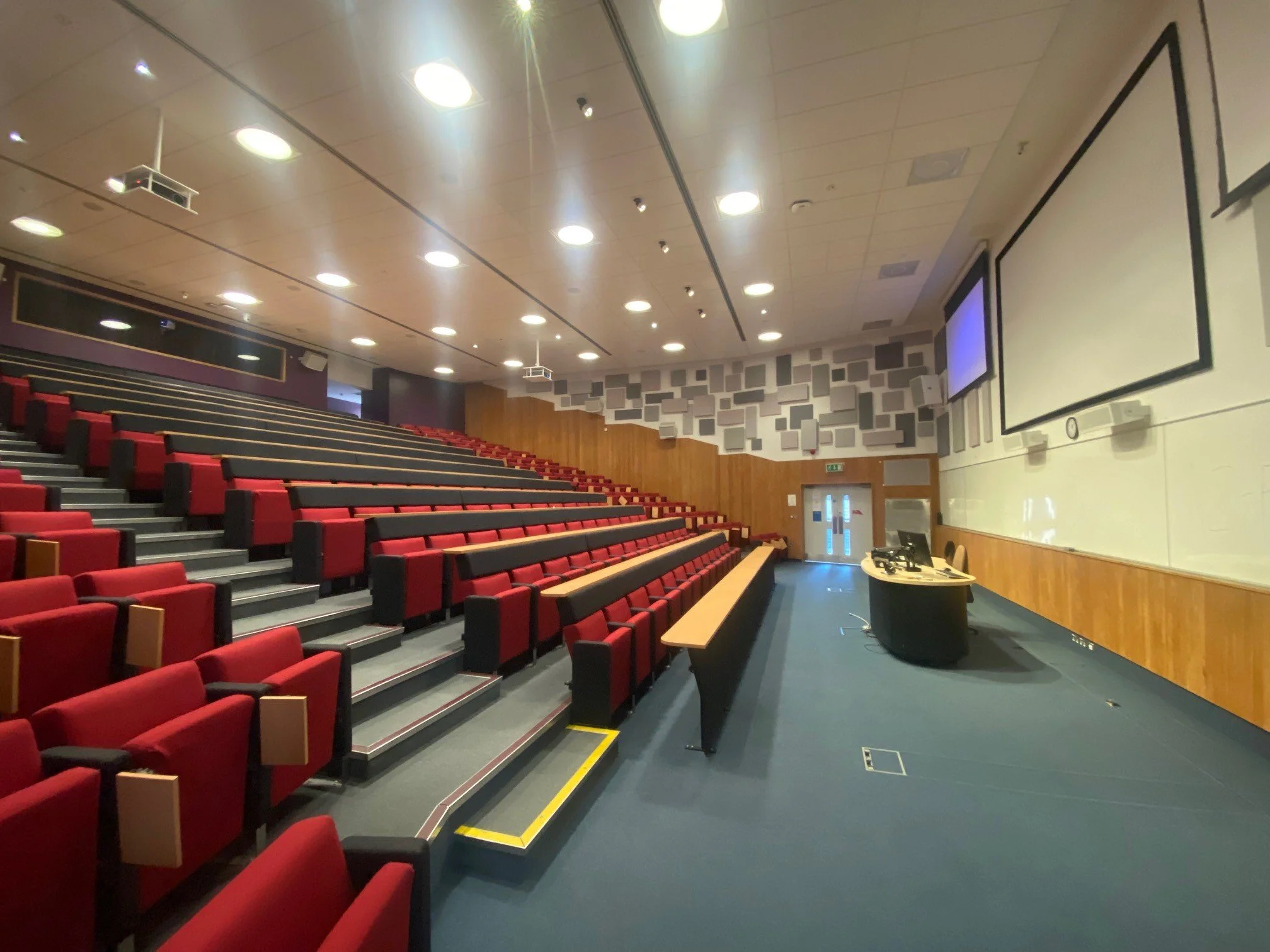 Empty lecture hall at the university of surrey with red cushioned seats, a large white projection screen, and a podium with a computer and microphone at the front.