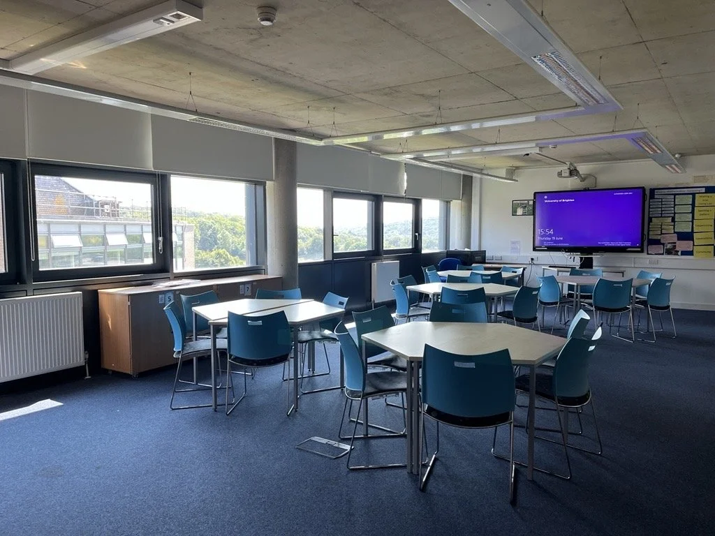University of Brighton classroom or meeting room with several white tables and blue chairs, large windows on one side, a digital display on the wall, and educational posters on the wall.