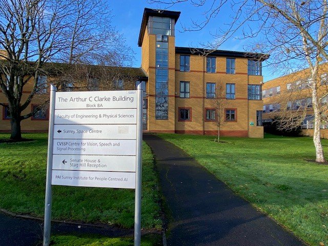 Signpost in front of a brick building at the university of surrey with multiple windows, on a grassy area with trees and a clear blue sky.