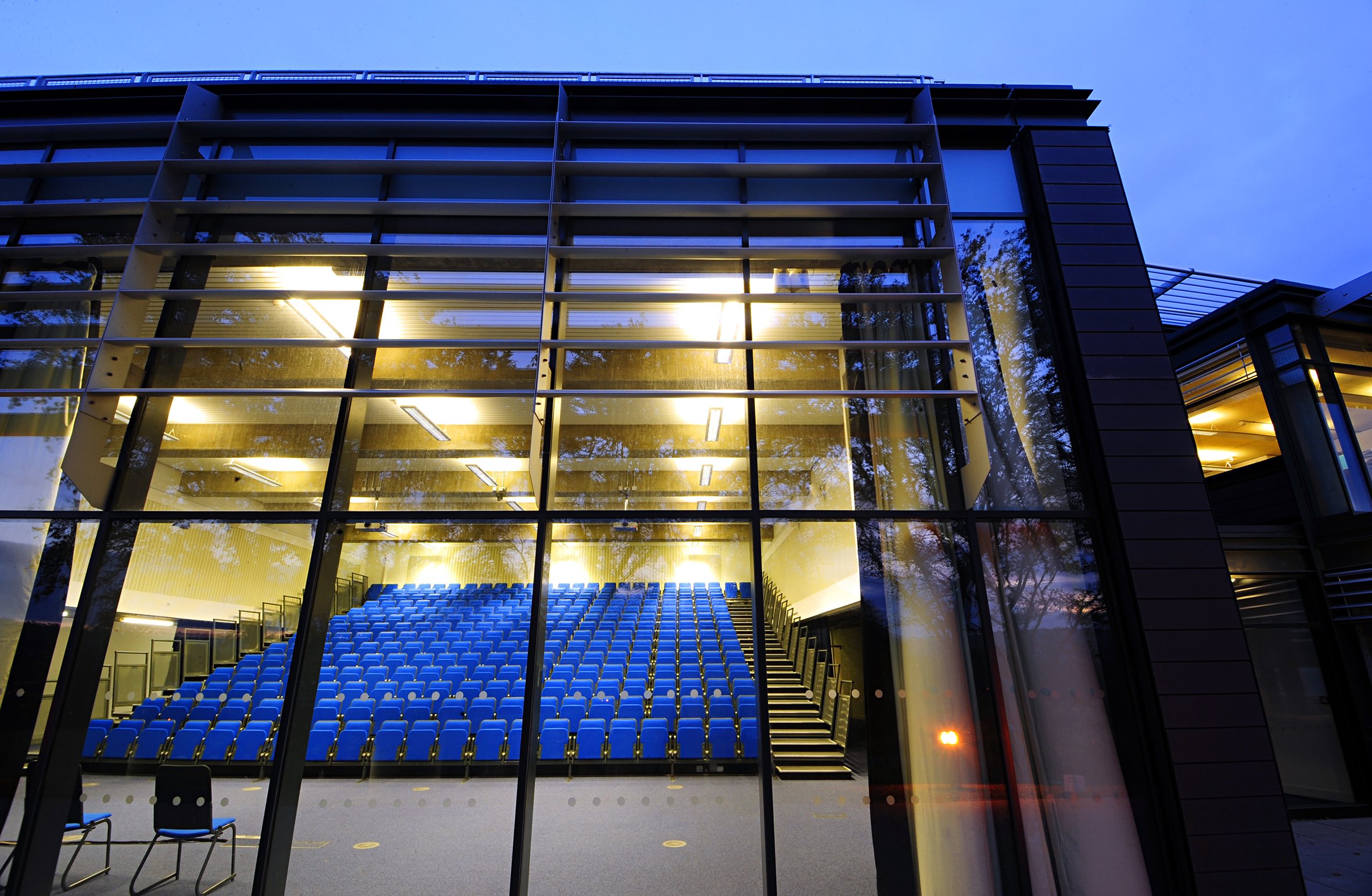 University of Brighton Exterior view of a modern building with large glass windows and blue seats inside, illuminated with warm lighting, taken during dusk.