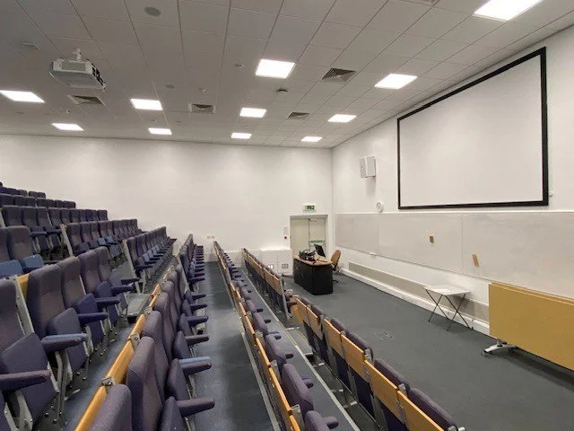 Lecture theatre at the university of surrey with rows of purple chairs, a large screen at the front, a podium with a microphone, and a small table.
