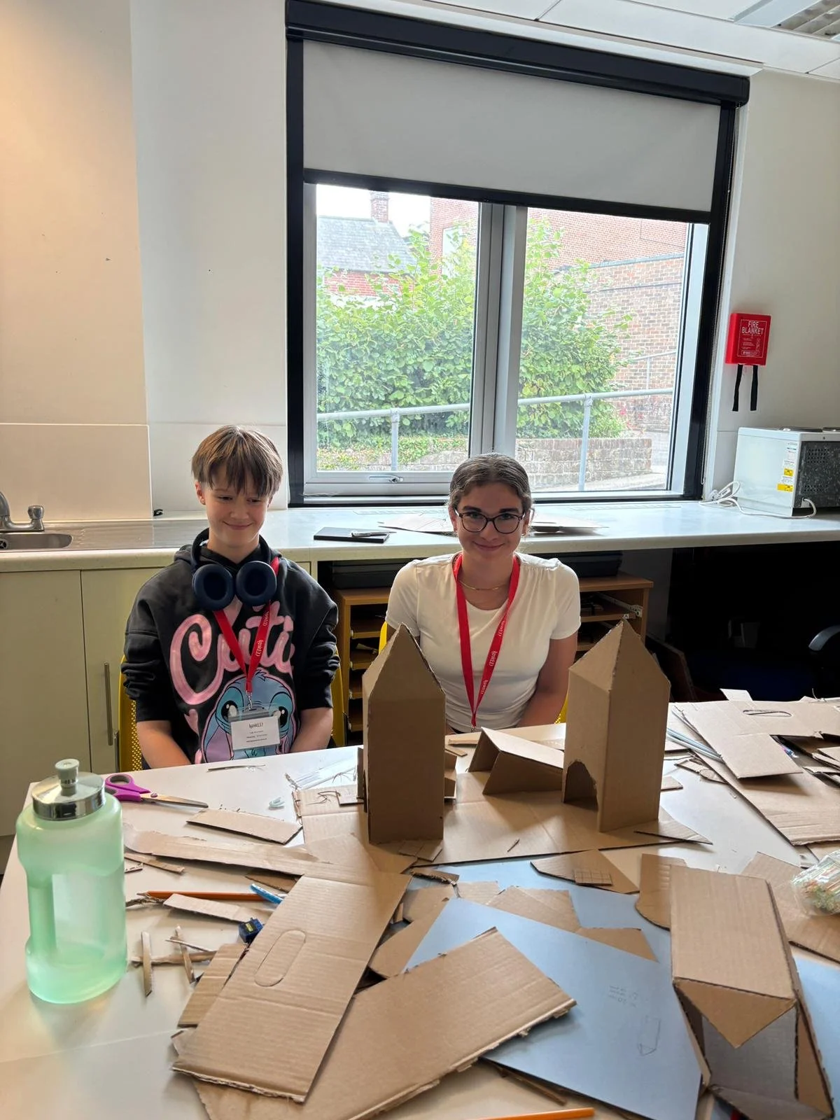 Two children sit at a table with cardboard structures and crafting supplies, in a room with a window showing greenery outside.
