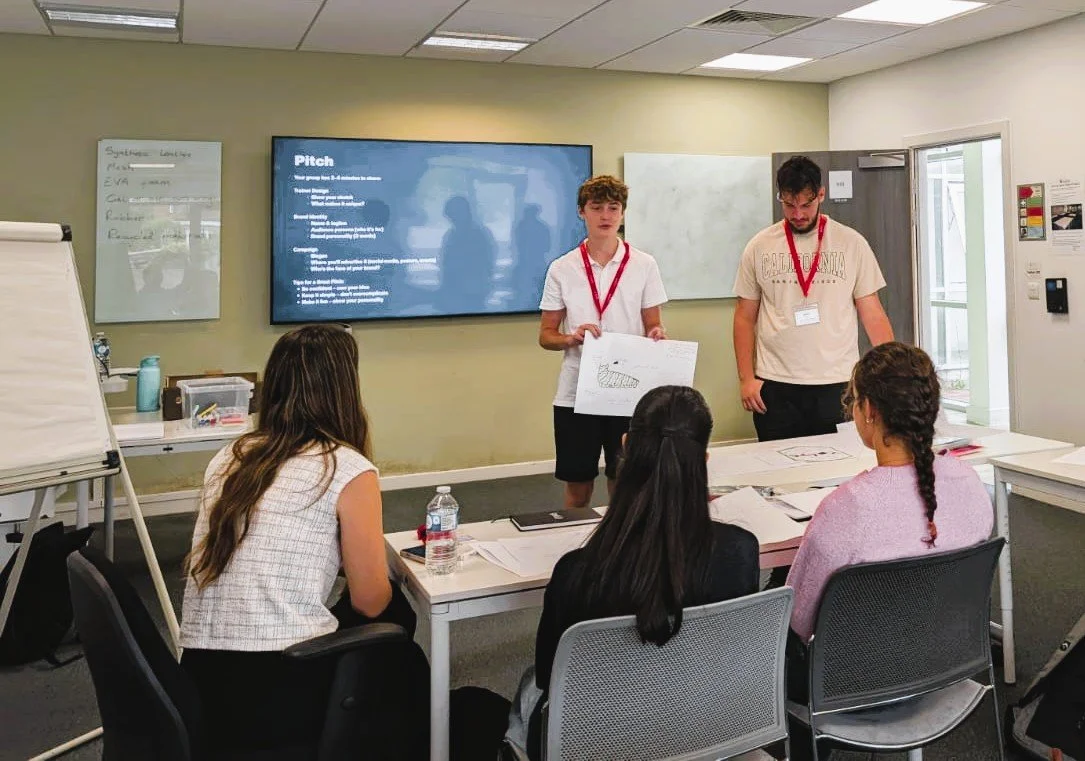 Two young men present a project to three seated women in a classroom. One man holds a drawing of a pie chart, and a large screen displays presentation notes.