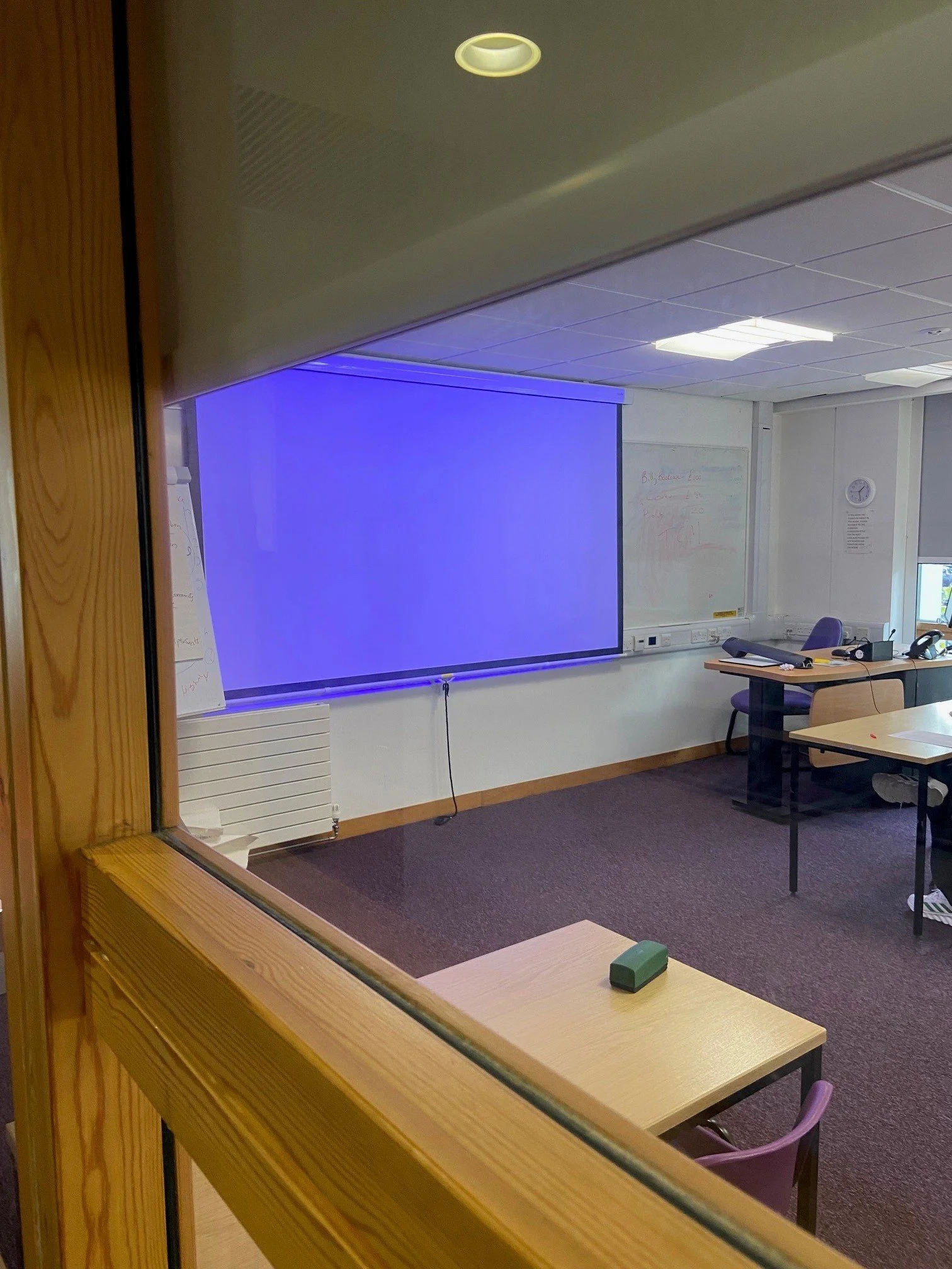 Empty classroom with a large blue screen or projection on the wall, whiteboards, and desks with chairs, viewed through a window or open door.