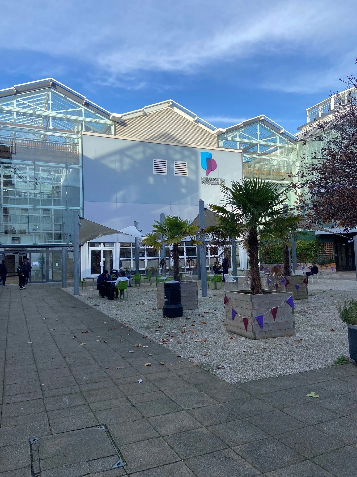 Exterior view of University of Portsmouth Forum building with glass structures, palm trees in planters, and outdoor seating area, under a partly cloudy sky.